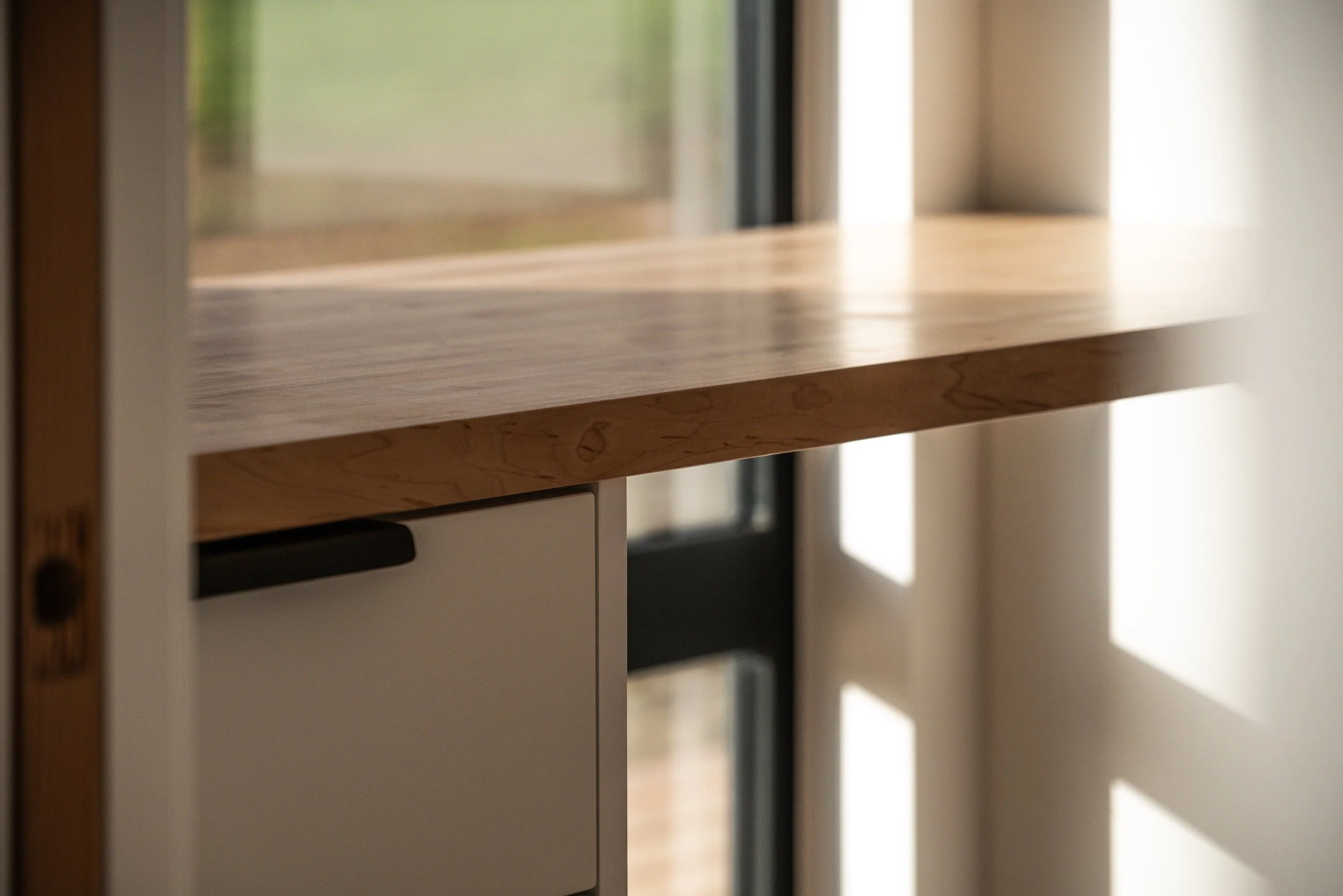 Close-up of a wooden countertop with natural wood grain, adjacent to a black-framed glass door and a white cabinet, with sunlight casting shadows on the wall.