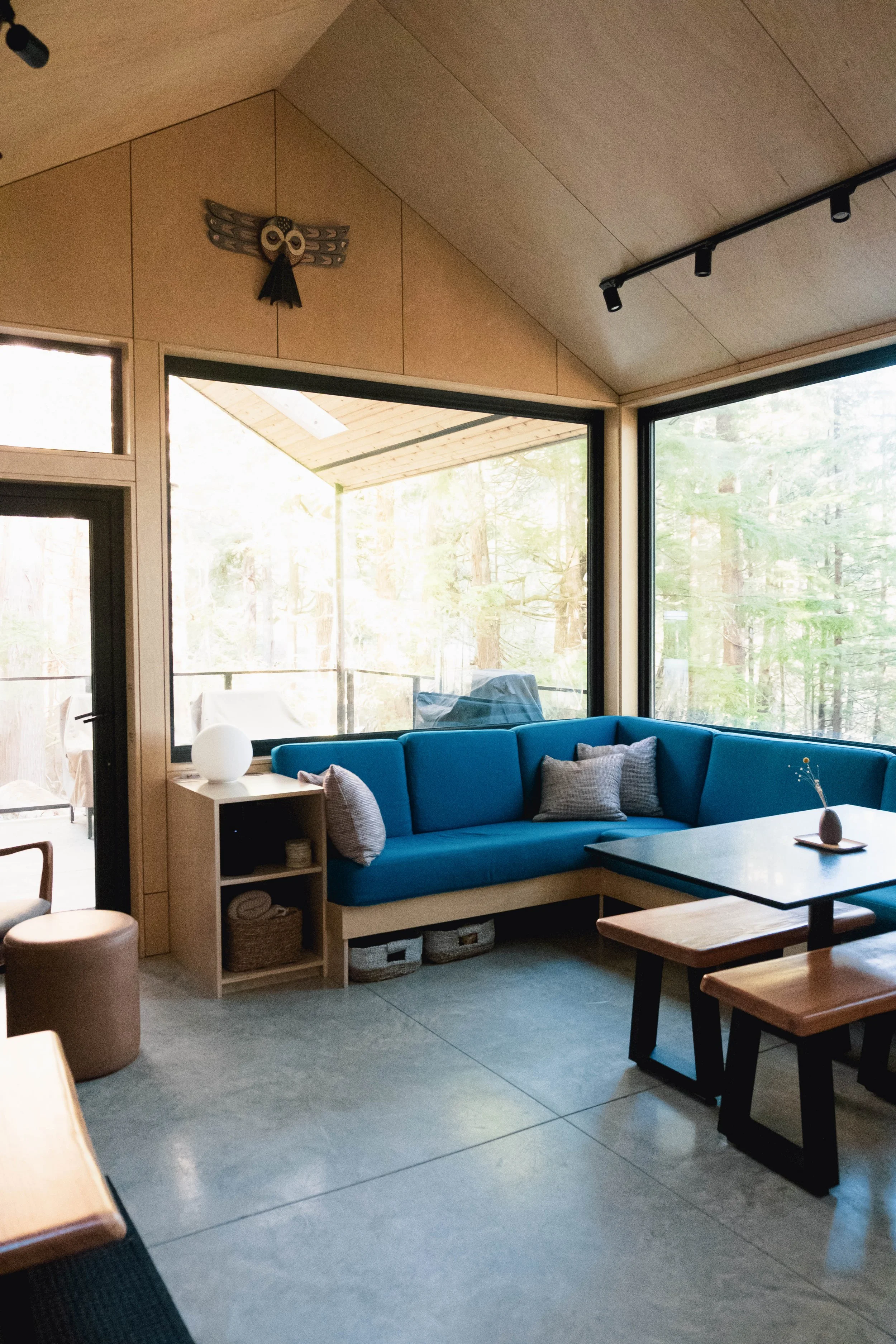 Living room with large windows, blue couch with pillows, black coffee table, wooden table with benches, and decorative owl wall art.