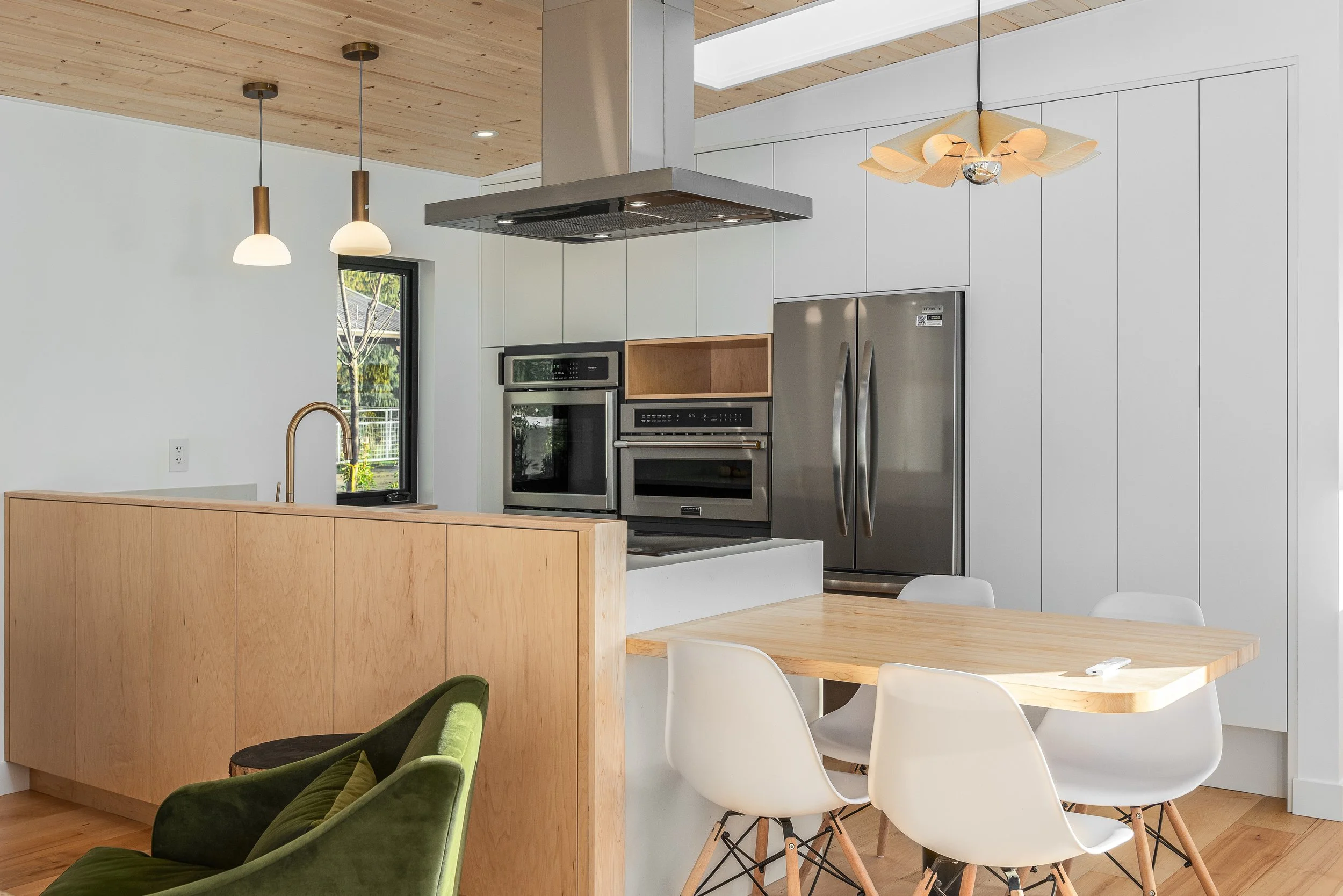 Modern kitchen with stainless steel appliances, white cabinets, a wooden dining table with white chairs, and a green chair in the foreground, natural light coming through a window.