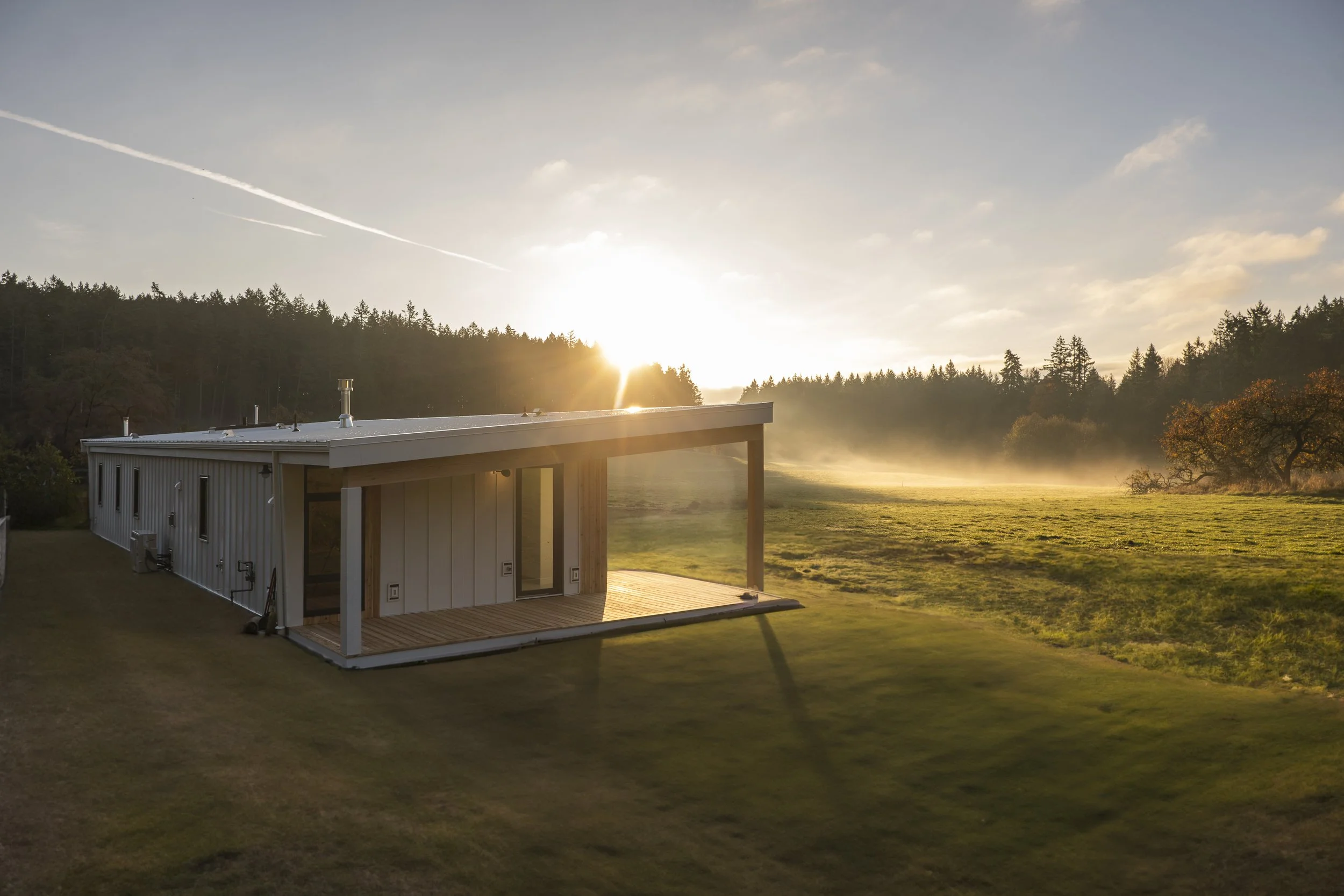 Modern white house with a wooden deck in a green field during sunrise, with mist in the background and trees on the horizon.