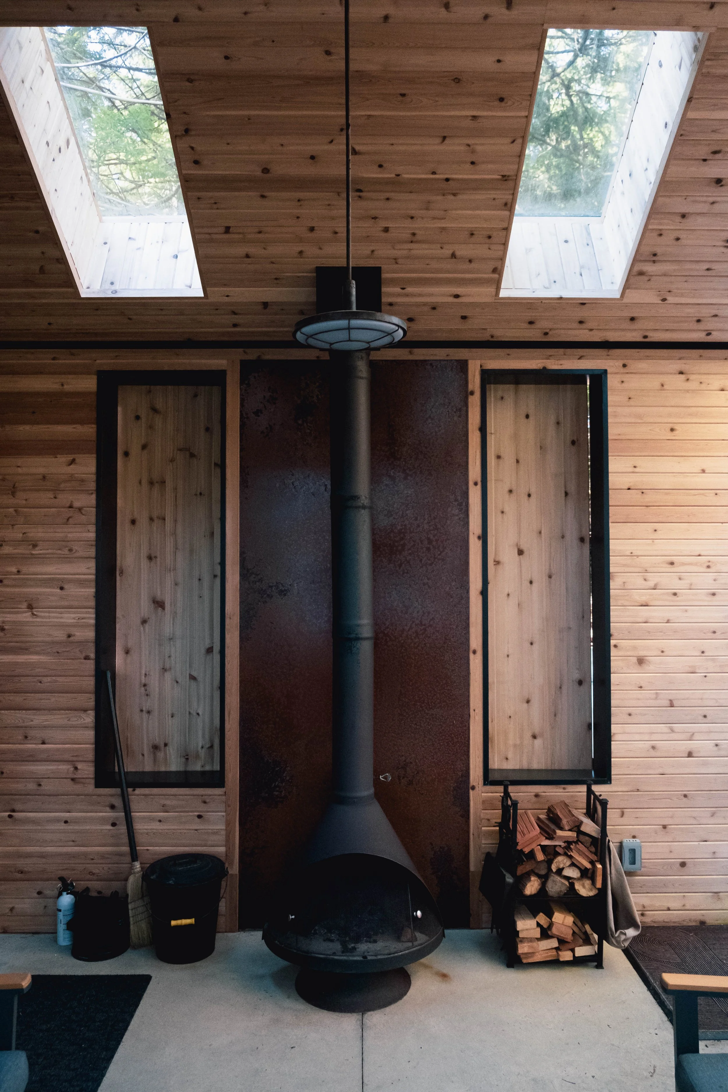A wood-paneled room with a central black wood stove with a chimney pipe extending upward, flanked by two windows in the ceiling, and a wood storage area on the right side.