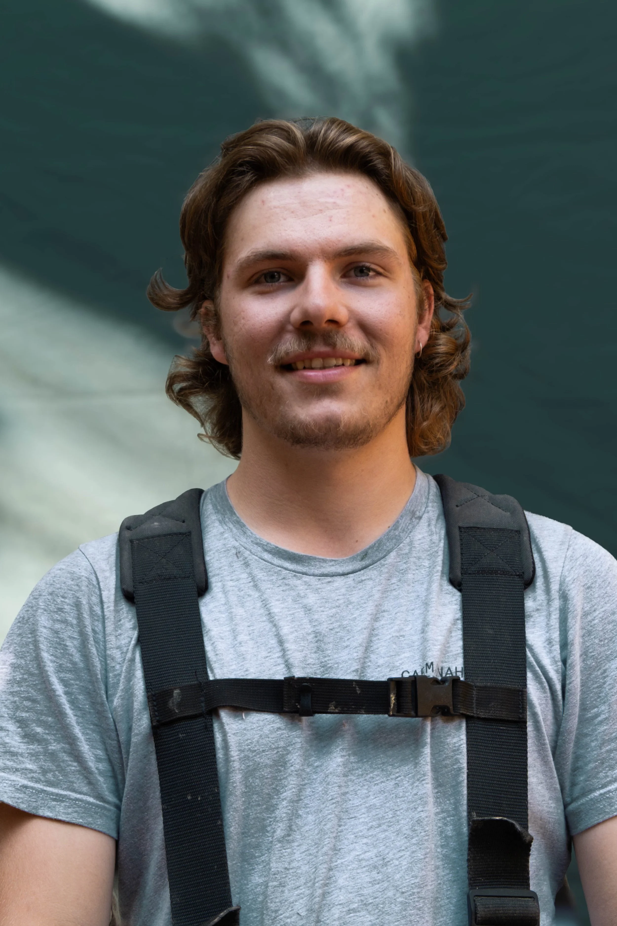 A young man with wavy brown hair, a mustache, and freckles, wearing a gray t-shirt with a black backpack, smiling in front of a blurred natural background.