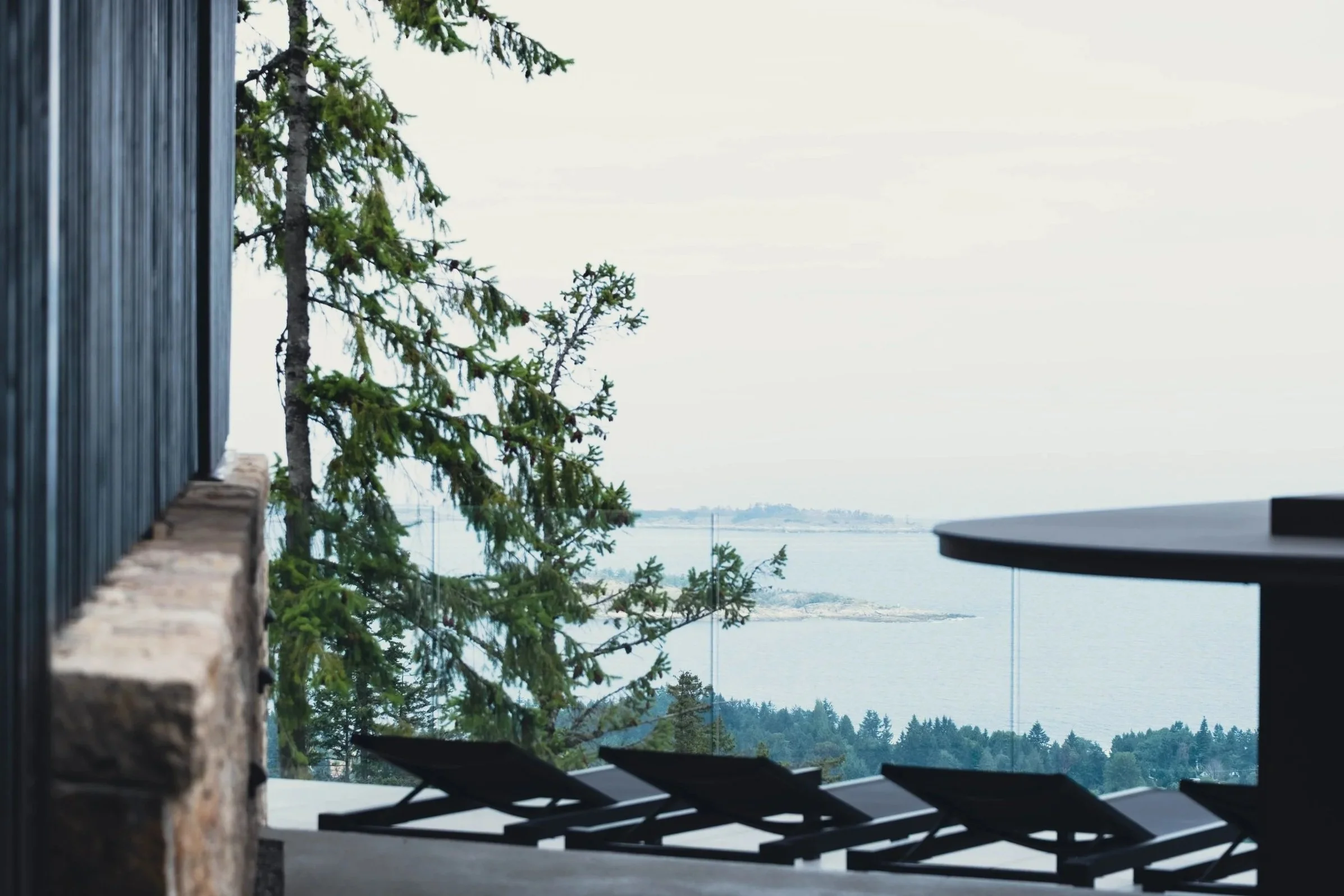 A scenic outdoor view of a lake with trees and shoreline in the distance, partially obstructed by a wall, with lounge chairs and a table in the foreground.
