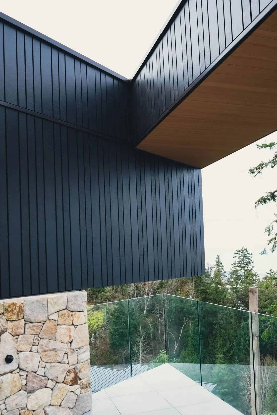 Modern building with dark vertical siding, a wooden ceiling overhang, stone wall, glass railing, and trees in the background.