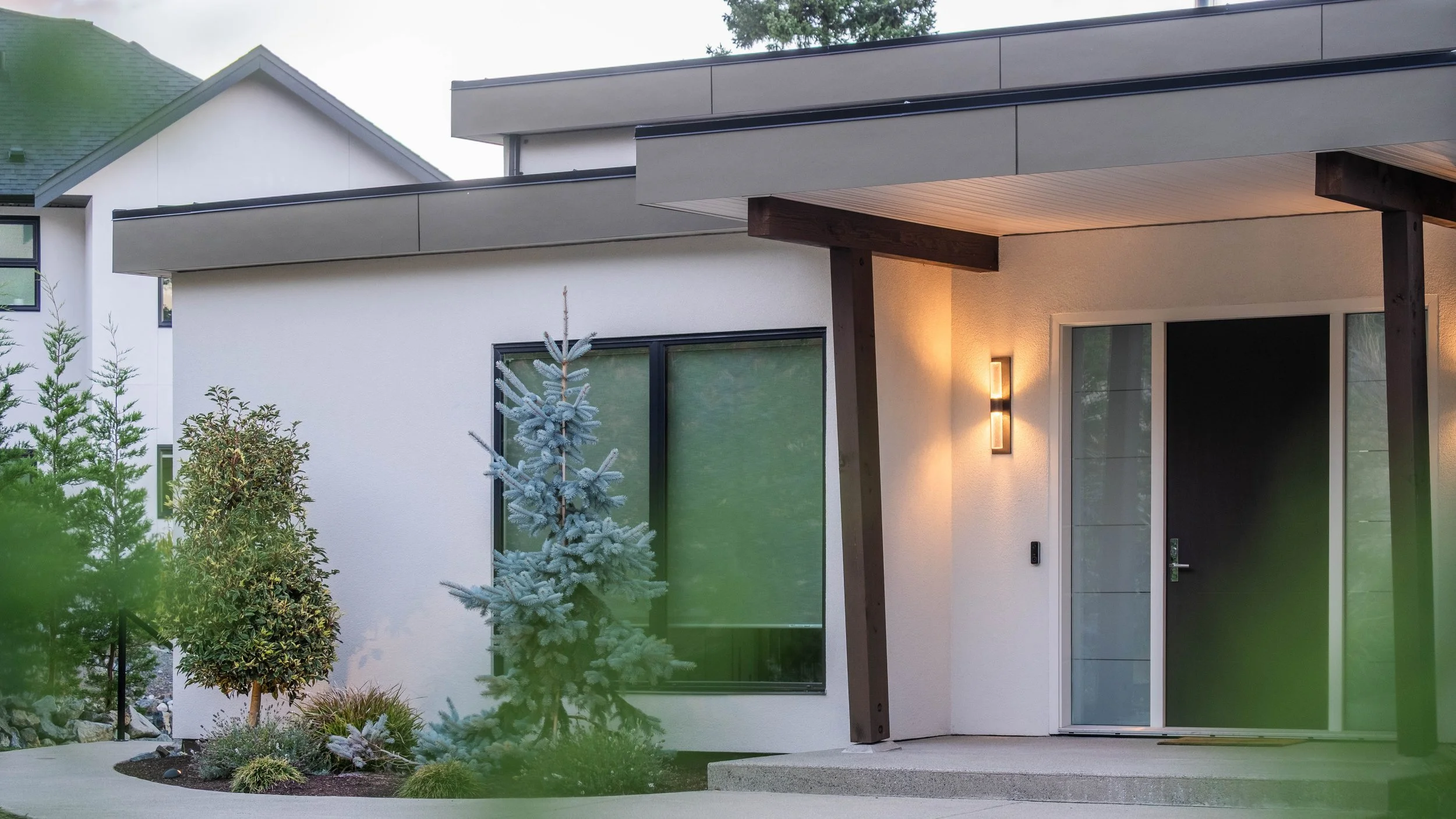 Modern house exterior with white walls, large window, sliding glass door, and outdoor lighting, surrounded by garden plants and small trees.