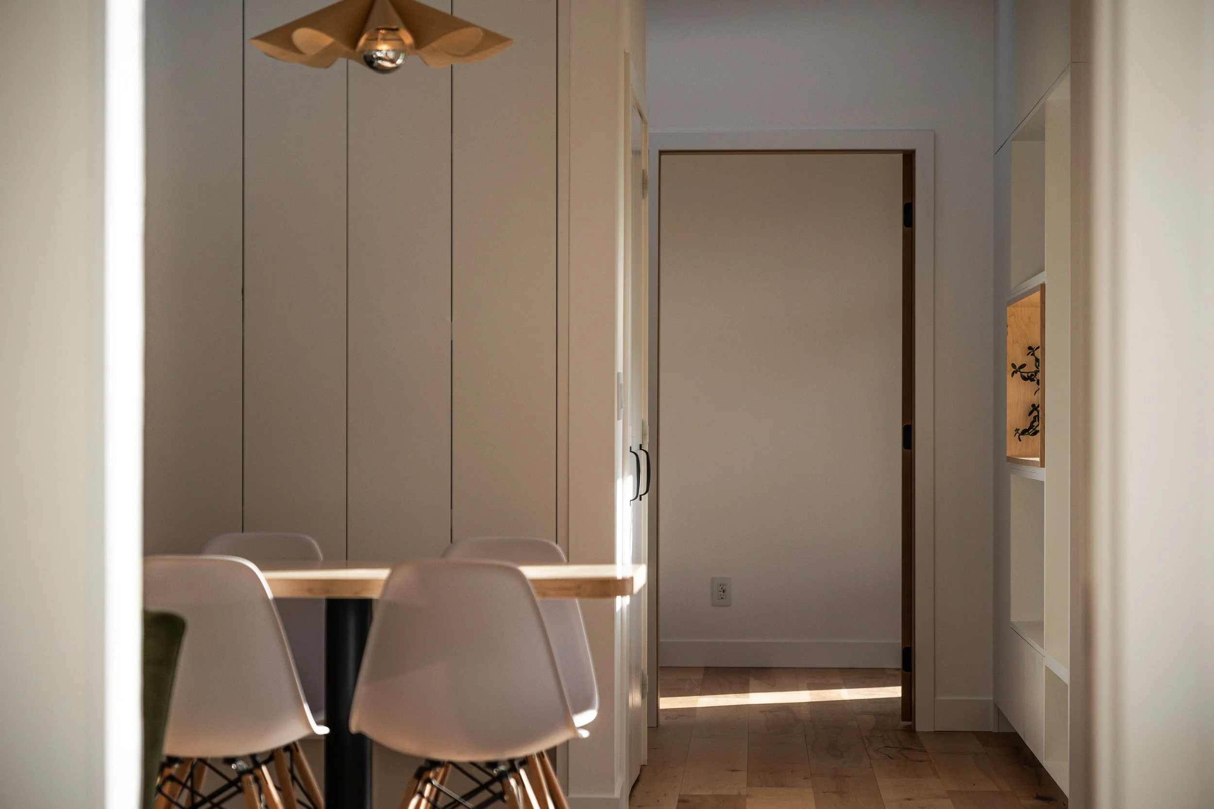 Interior view of a modern kitchen or dining area with white cabinets, a wooden table, white chairs, and a pendant ceiling light. There is a doorway leading to another room and built-in shelves on the side.
