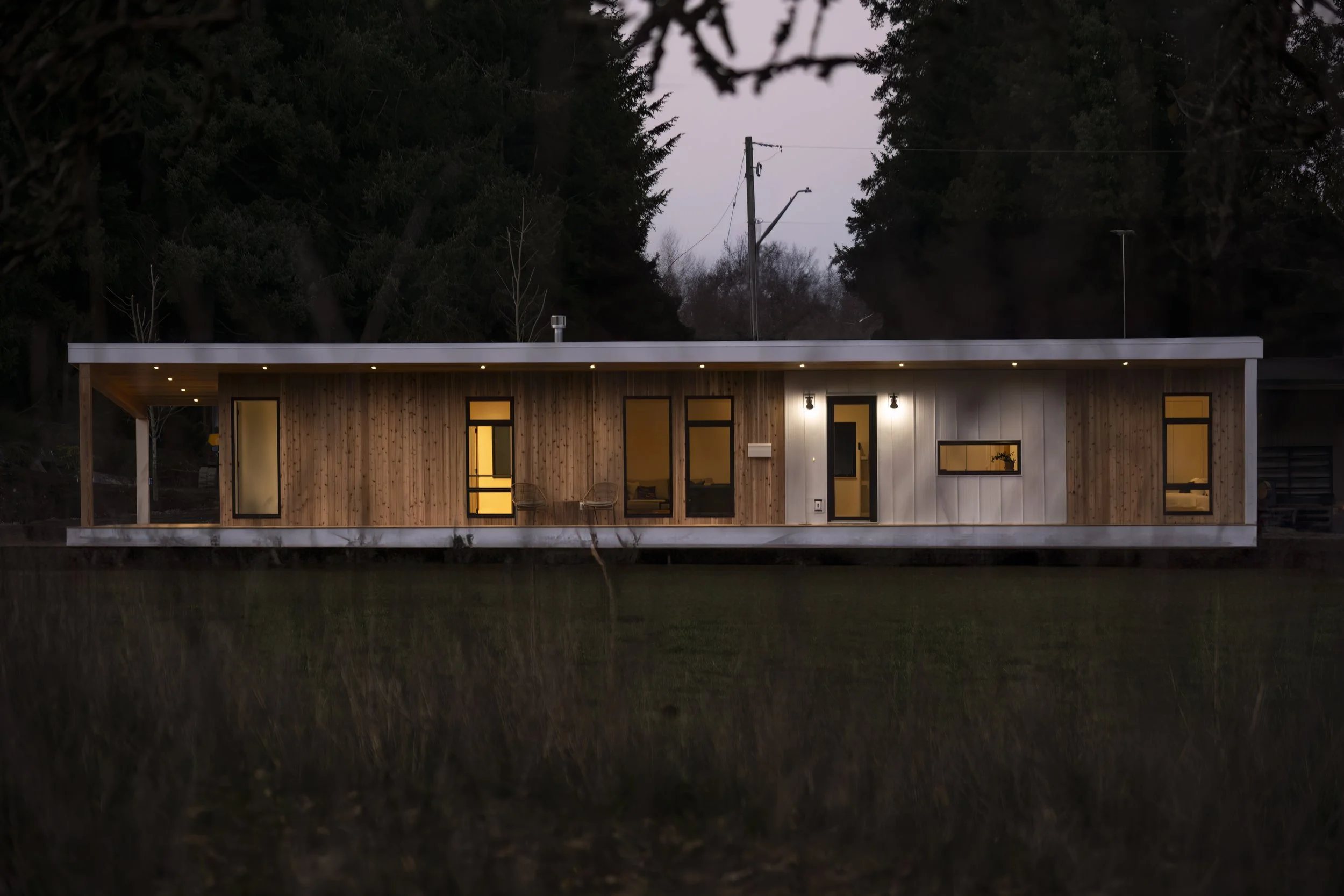Modern tiny house with wooden exterior, large windows, and outdoor seating, illuminated at dusk, situated near water with trees in the background.