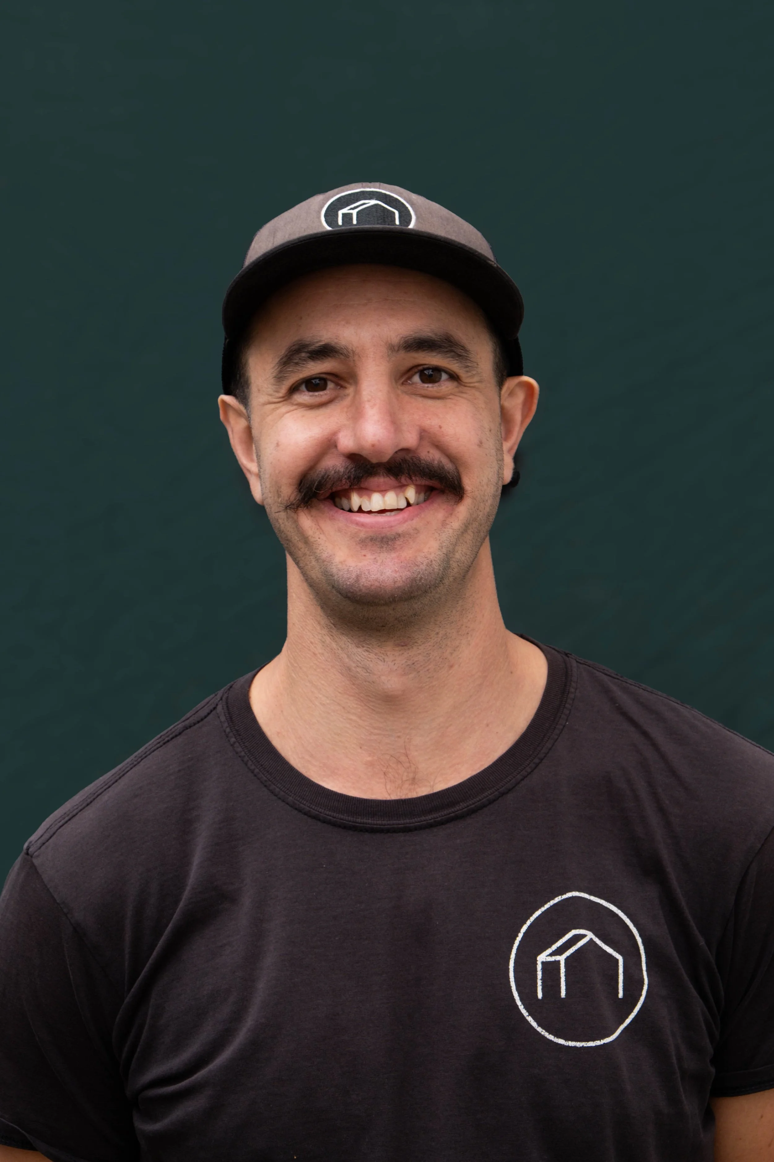 A smiling man wearing a black T-shirt and a gray baseball cap standing outdoors against a green background.