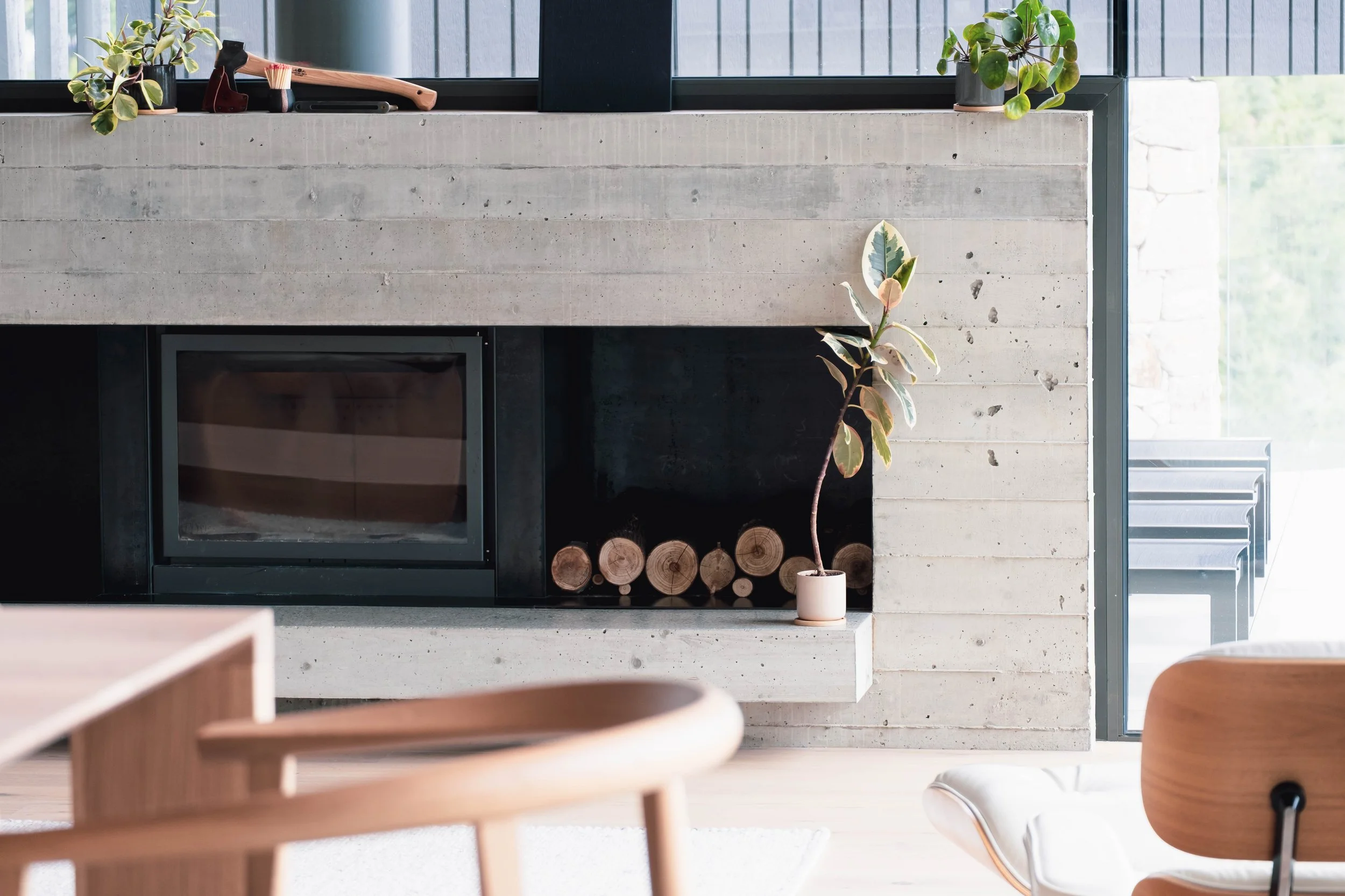 Modern living room with a concrete fireplace, potted plants on the shelf, and a stack of logs inside the fireplace. A large window with sunlight shining through.