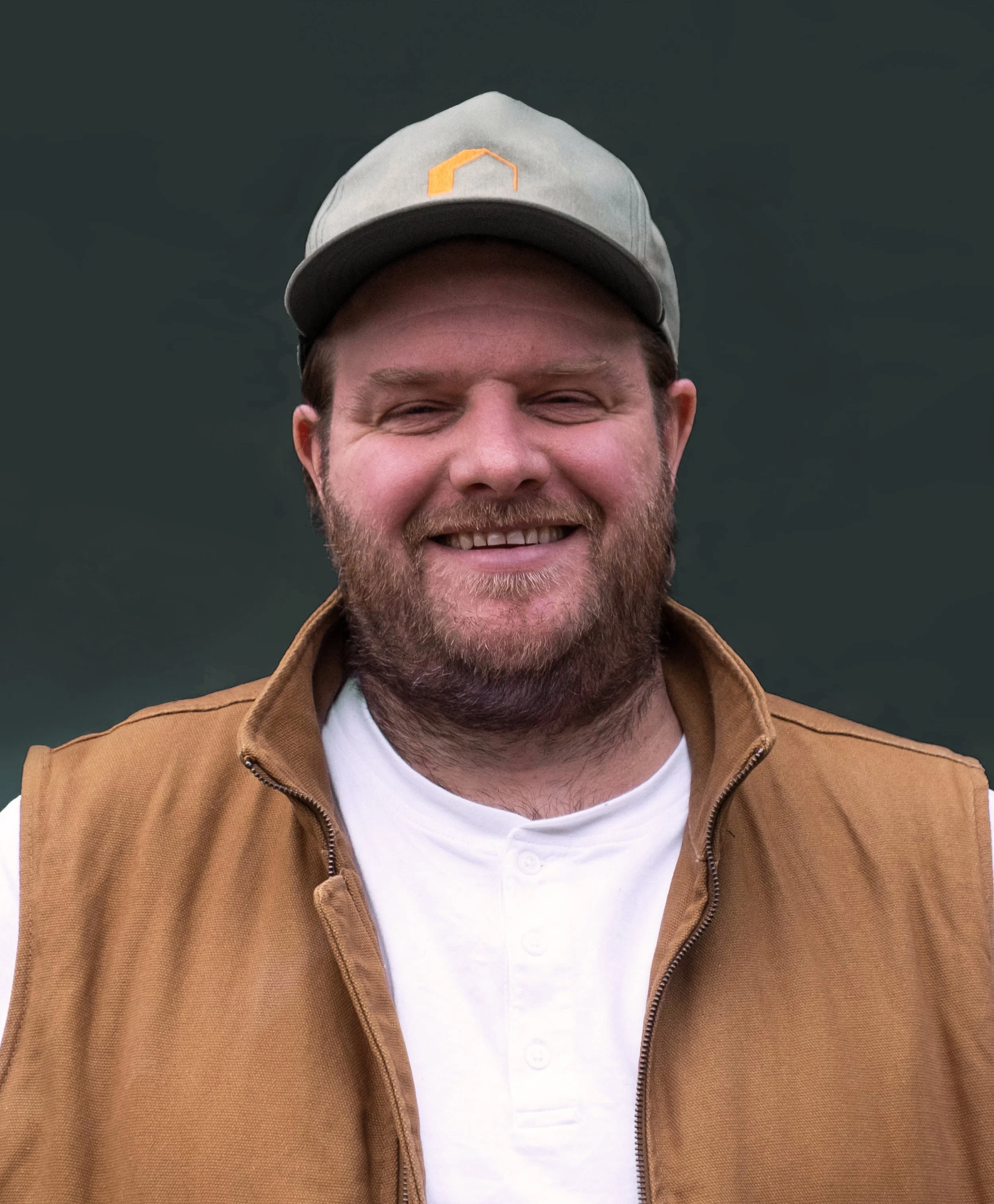 A smiling man with a beard wearing a gray cap and a brown vest over a white shirt, with a dark background.