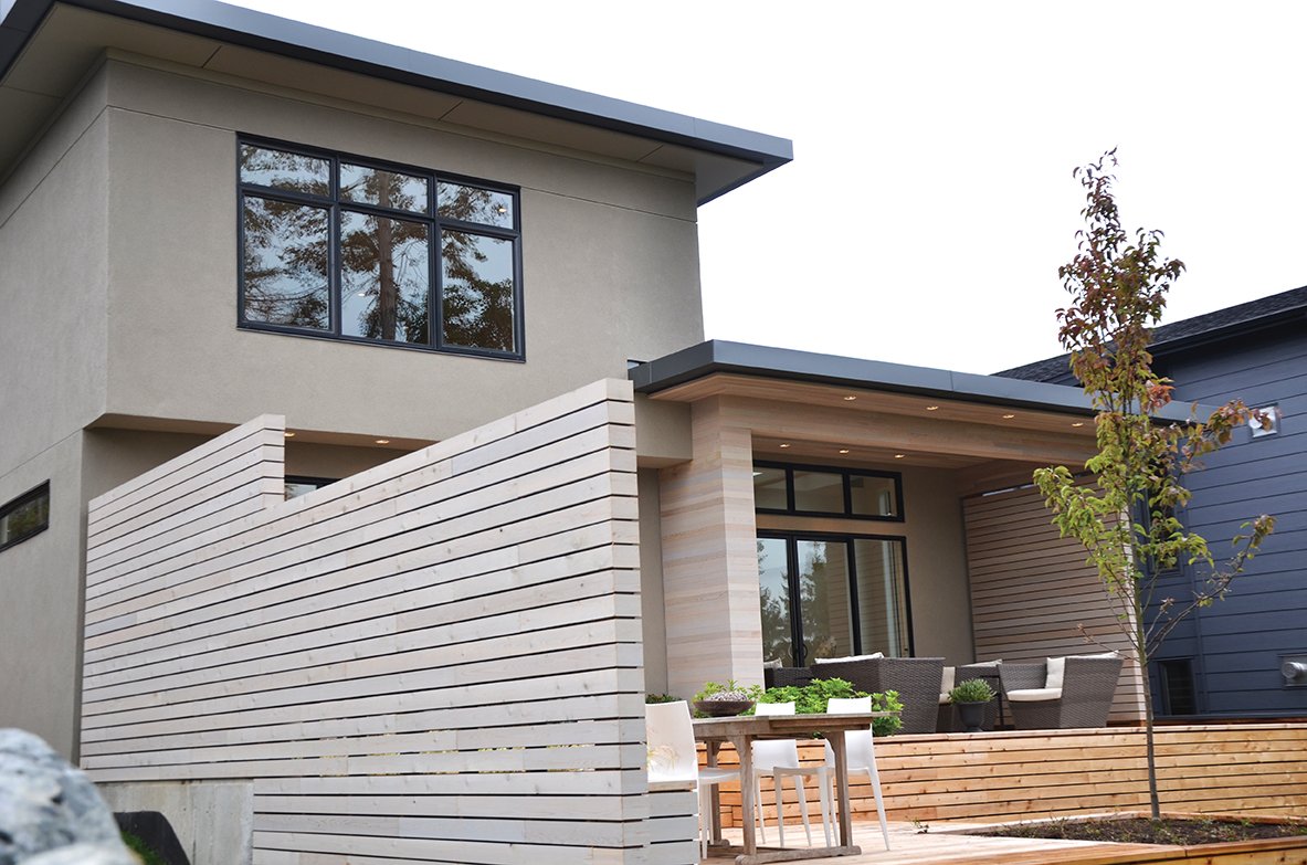 Modern two-story house with beige and blue exterior, large windows, and wooden patio with outdoor seating, trees, and a privacy fence.