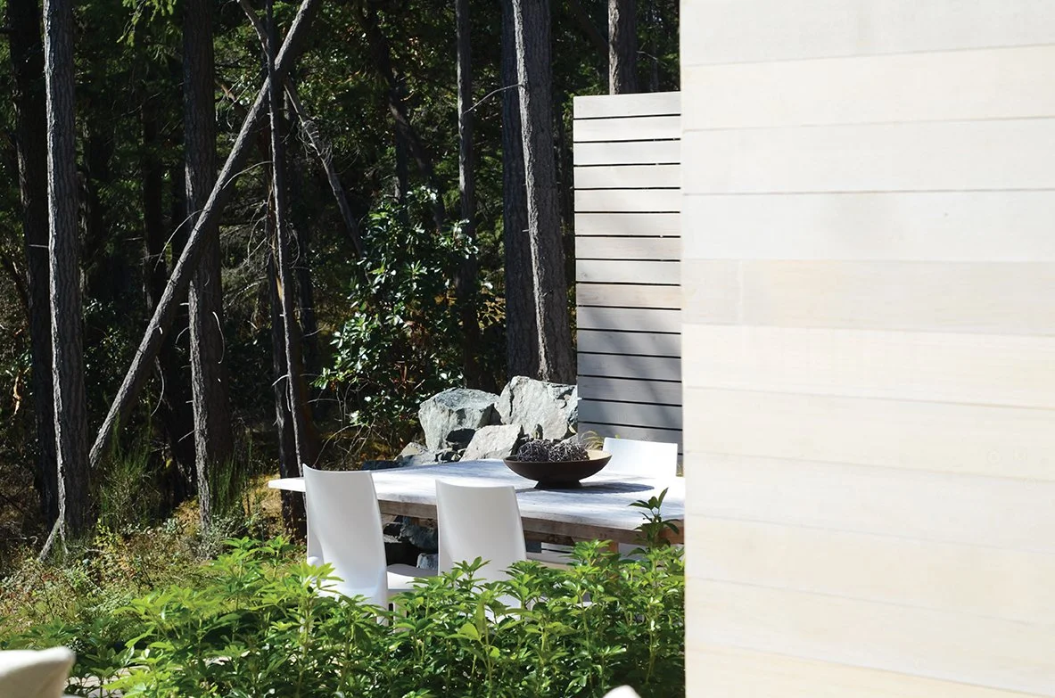 Outdoor dining area with a white table and white chairs, surrounded by greenery and trees, partially obscured by a beige wall.