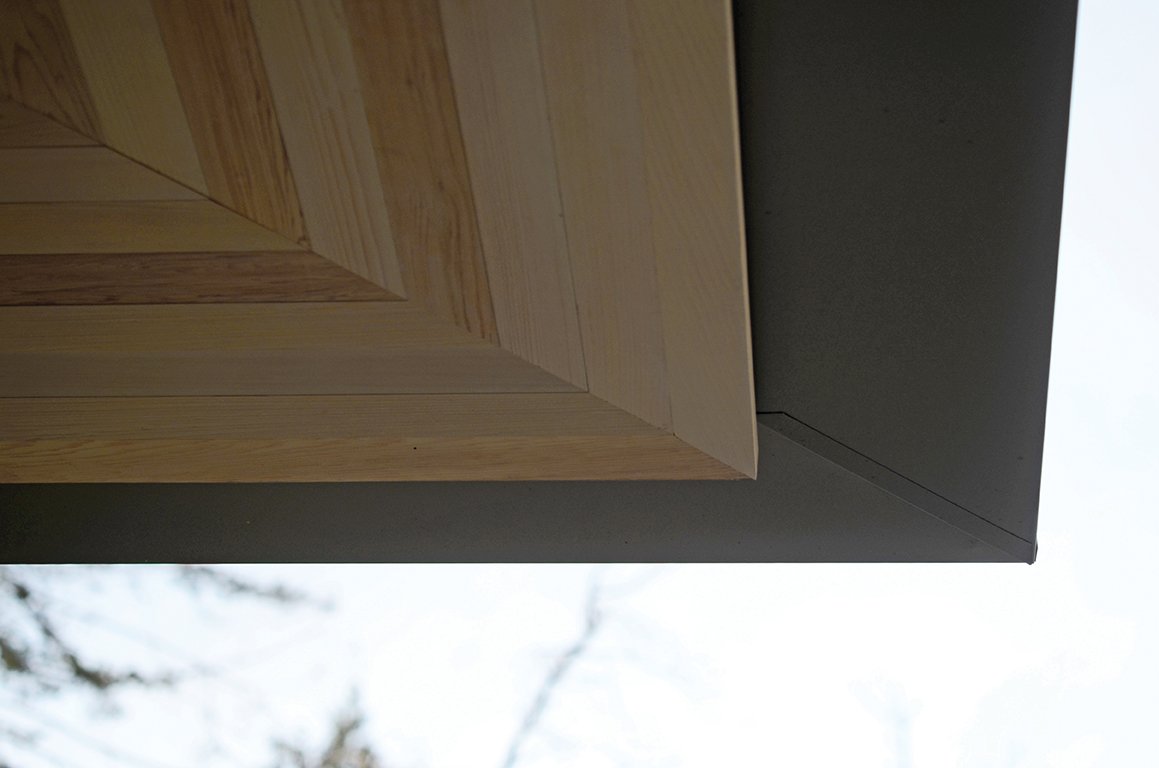 Close-up of a corner of a modern outdoor structure with wooden and metal elements, with trees and sky in the background.