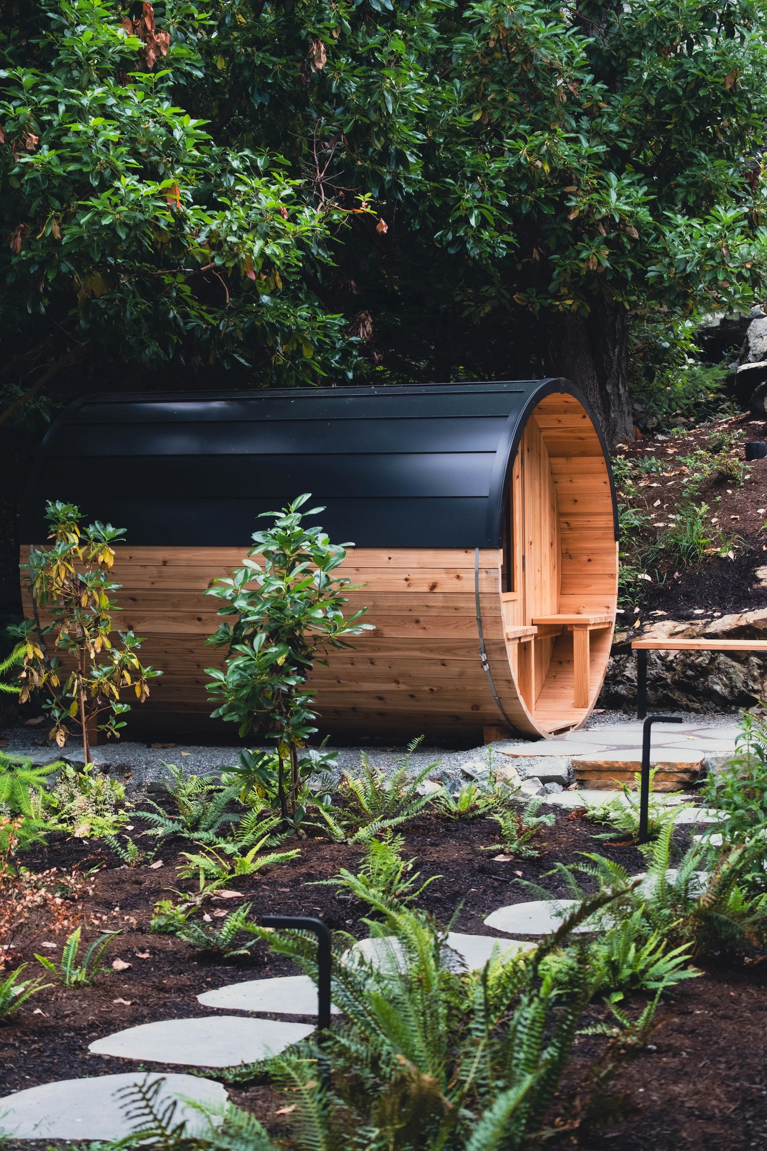 A small, cylindrical wooden sauna with a black roof, situated outdoors among lush green plants and trees, with a stone pathway leading to its entrance.