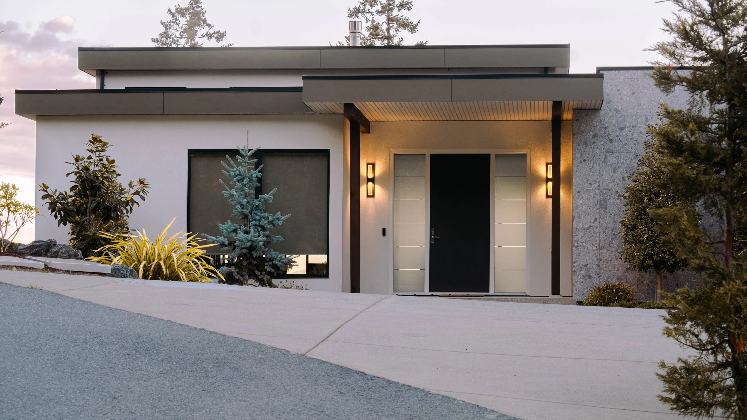 Modern house with a flat roof, front door, wall-mounted lights, and landscaped front yard with trees and plants.