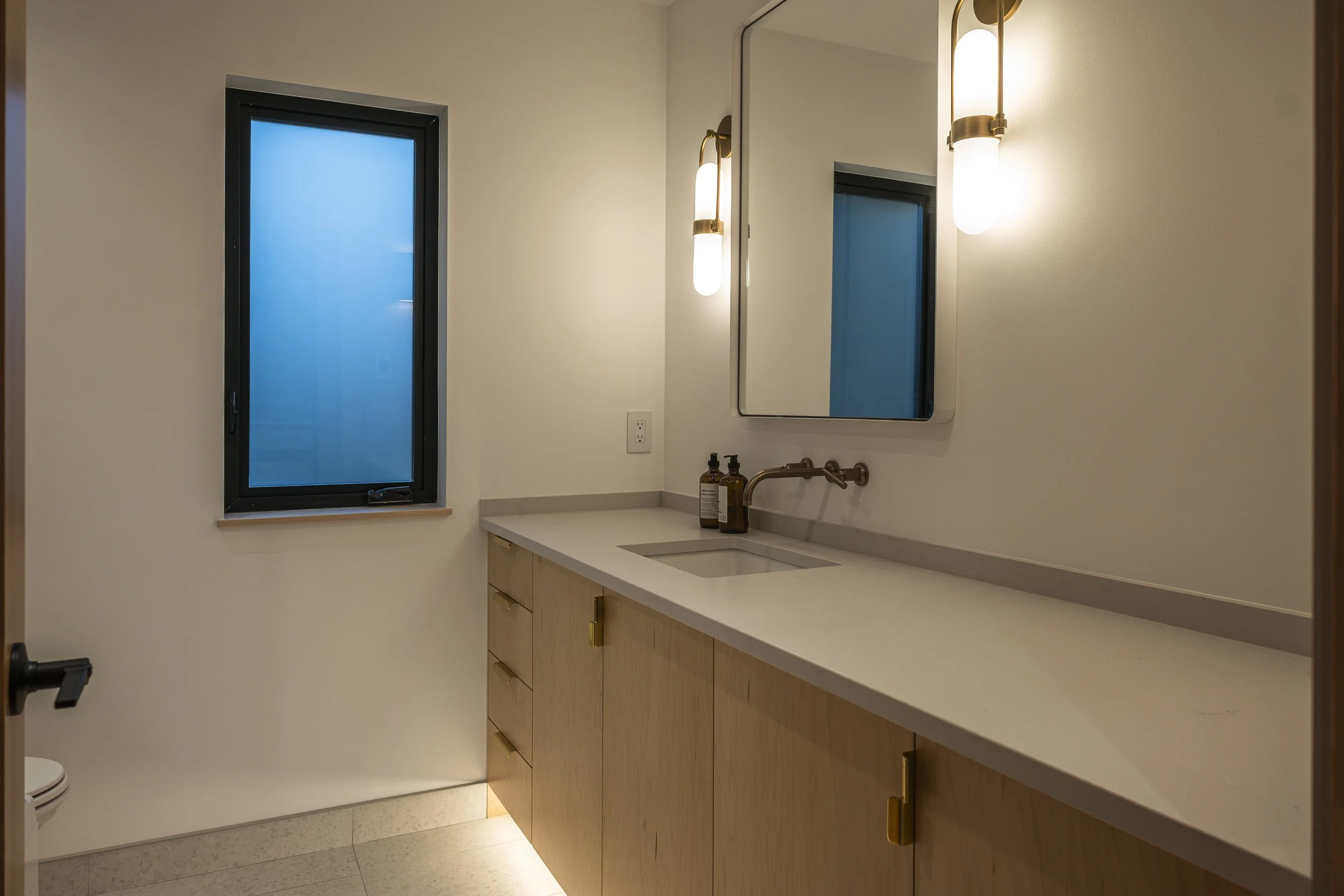 Modern bathroom with a wooden vanity, a mirror, two wall-mounted lights, a small window, and a sink with brown soap and lotion bottles.