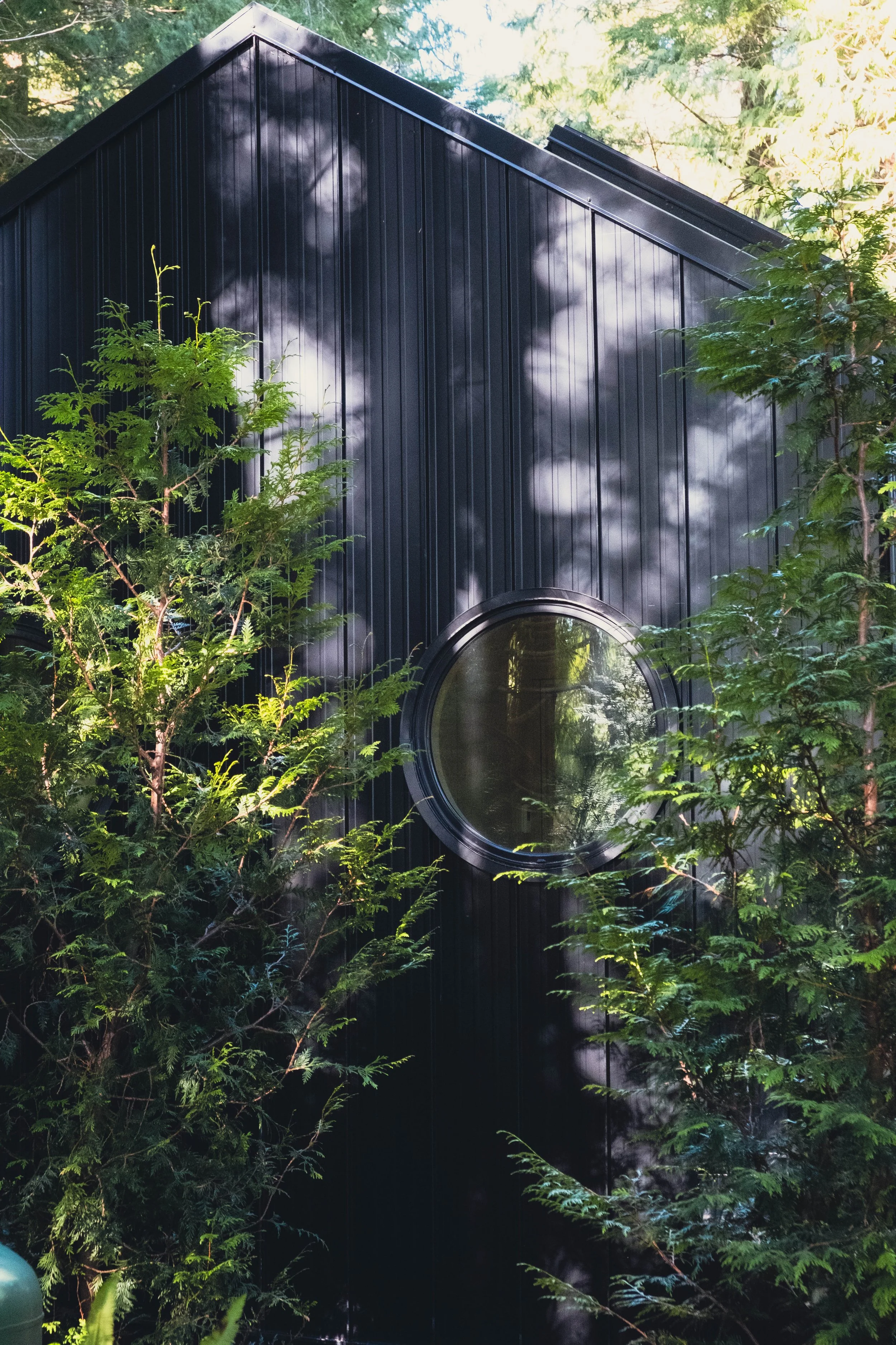 A black building with vertical siding and a circular window, surrounded by green trees.