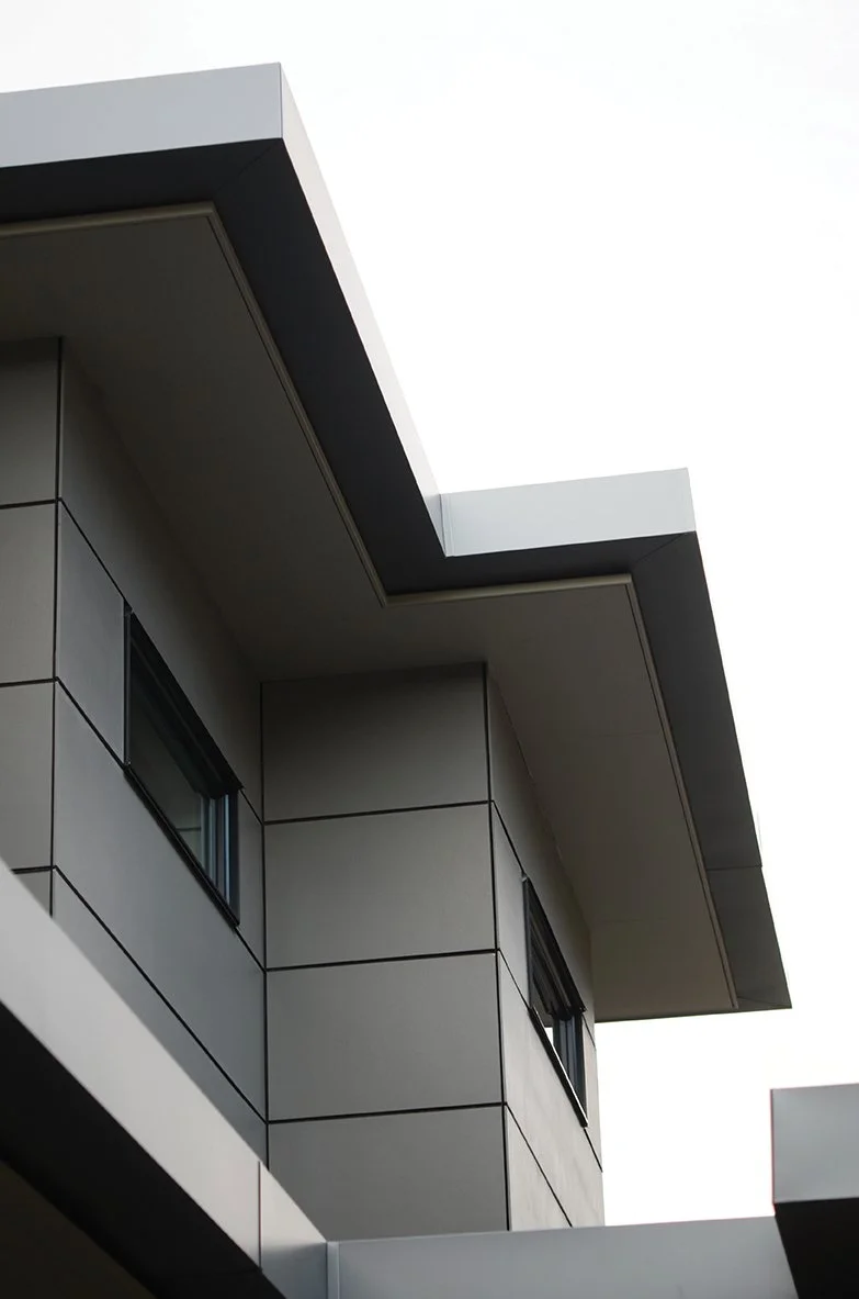 Close-up of a modern building exterior with beige panels, black window frames, and a protruding white roof overhang.