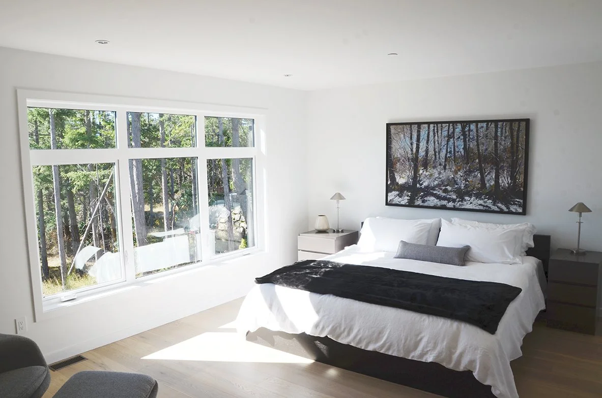 Bright bedroom with a large window showing a forest view, a bed with white bedding and gray pillows, two gray nightstands with lamps, and a framed landscape photograph of a snowy forest hanging above the bed.