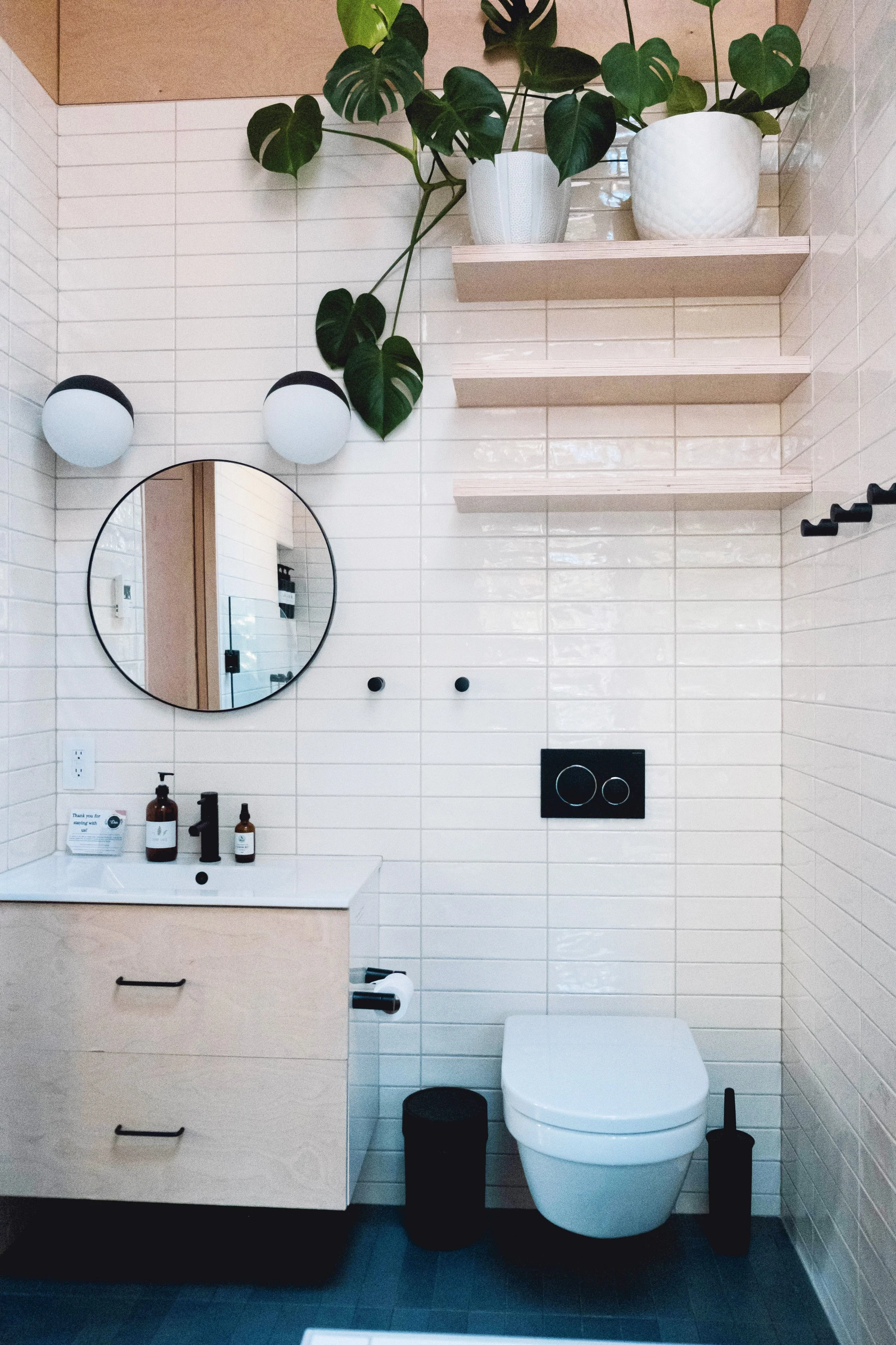 A modern bathroom with white tiled walls, a wooden vanity with black handles, a round mirror, decorative black light fixtures, black toilet paper holder, black trash can, and a white wall-mounted toilet. There are two floating shelves on the wall and