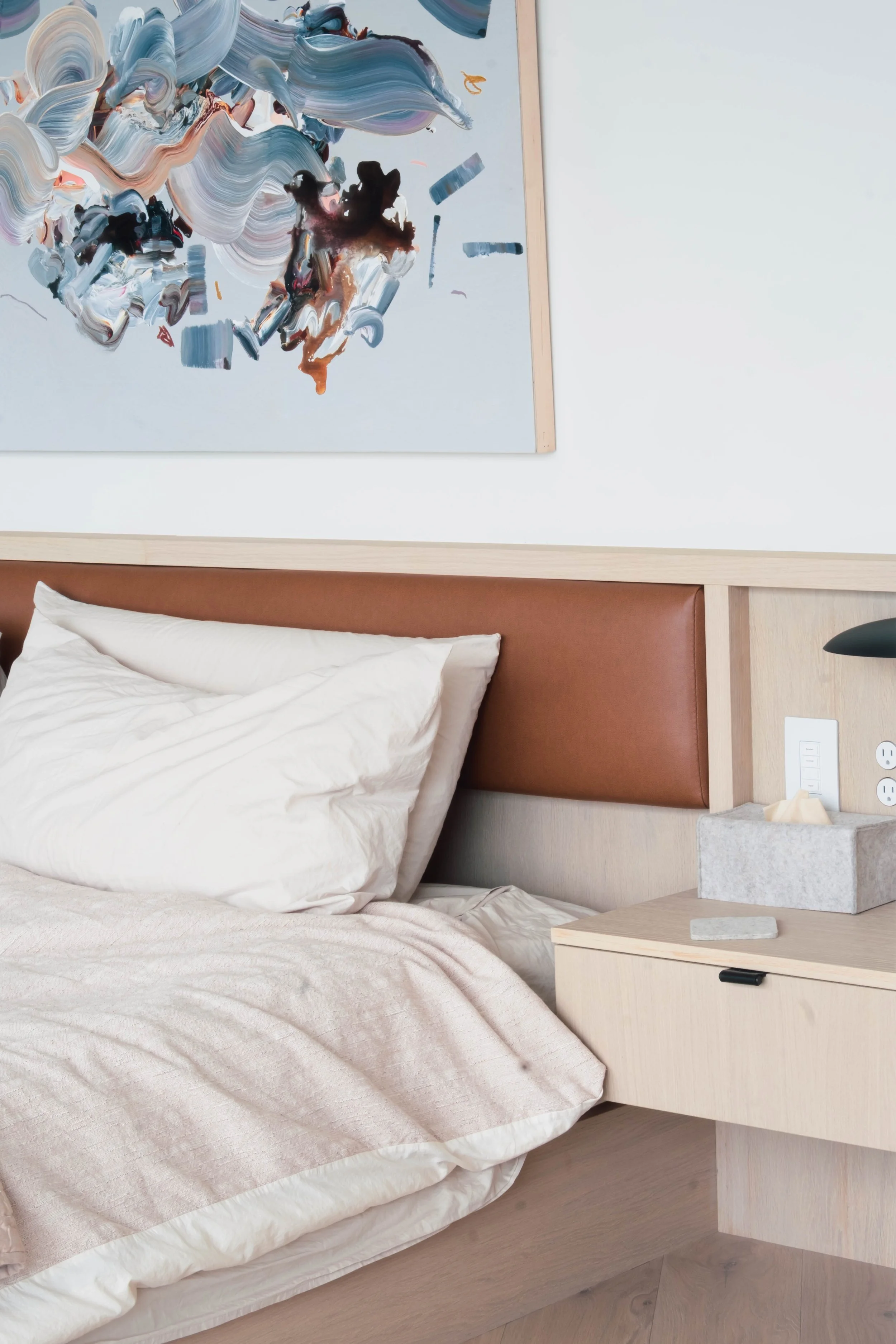 A neatly made bed with a white pillow and beige blanket, a wooden headboard with a brown leather section, a light wood nightstand with a tissue box and a small stone, and a modern abstract painting hanging on the white wall above the bed.