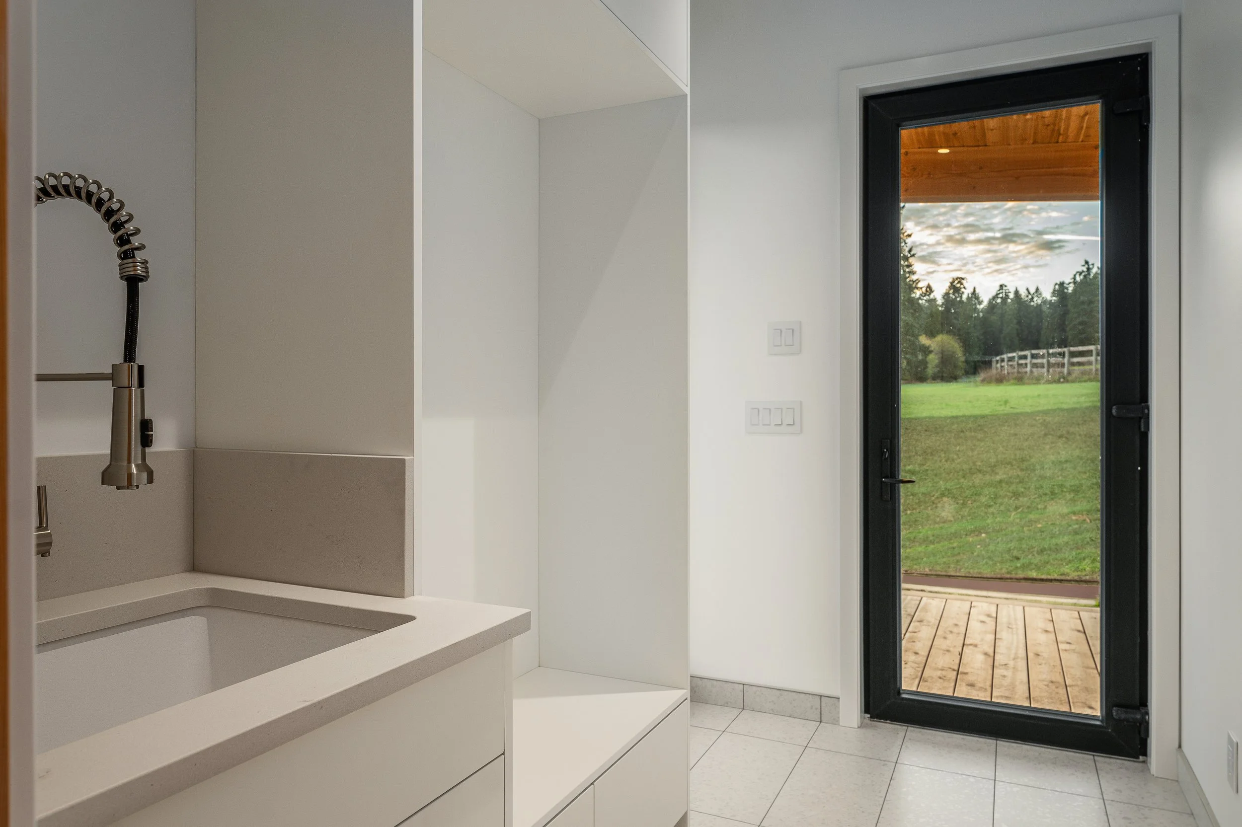 Modern interior of a room with a white sink, a black-framed glass door leading to an outdoor deck, and a scenic view of a green lawn and trees outside.