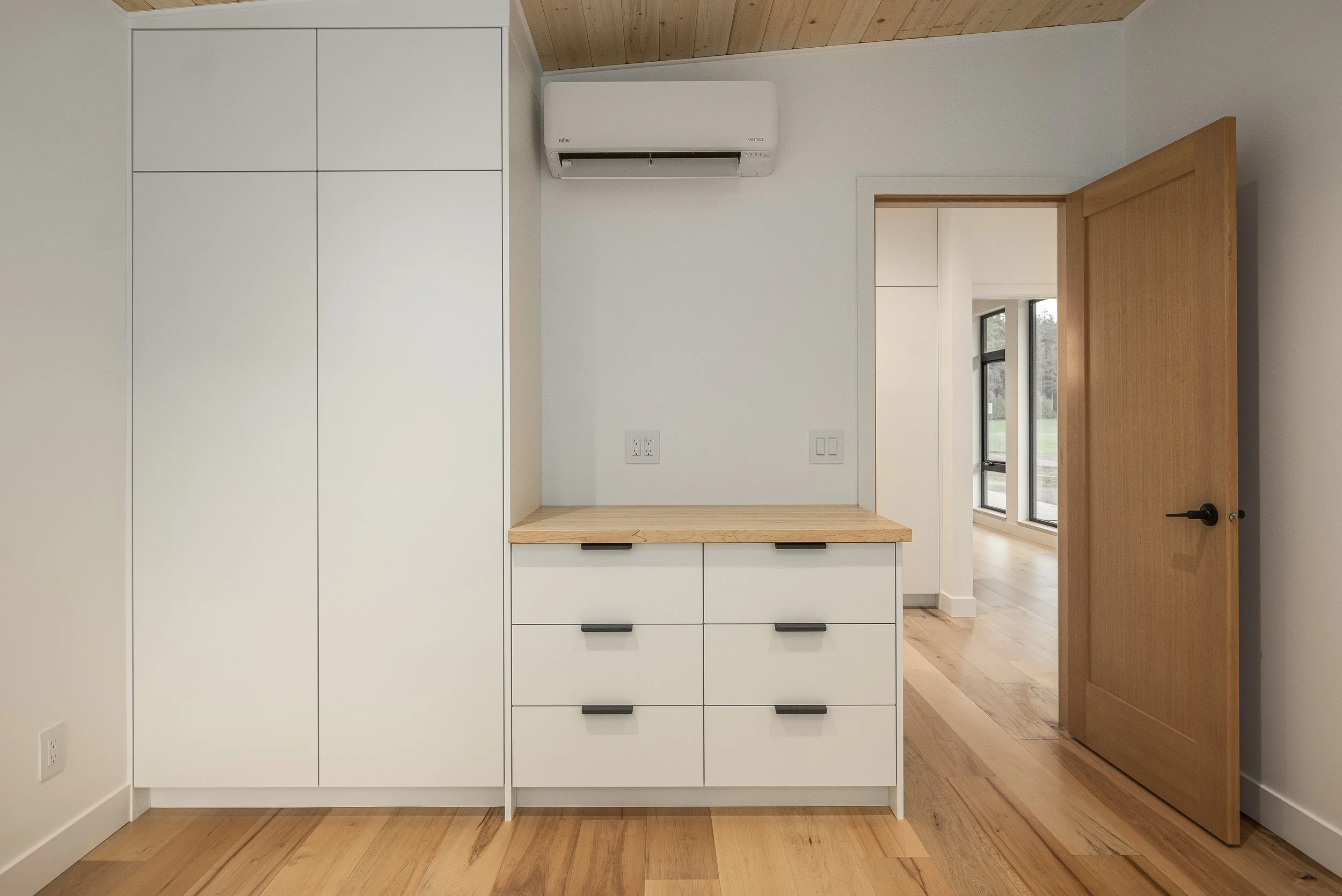 Interior of a modern room with a white built-in cabinet, a wooden countertop, and an open wooden door leading to a sunlit room with large windows.