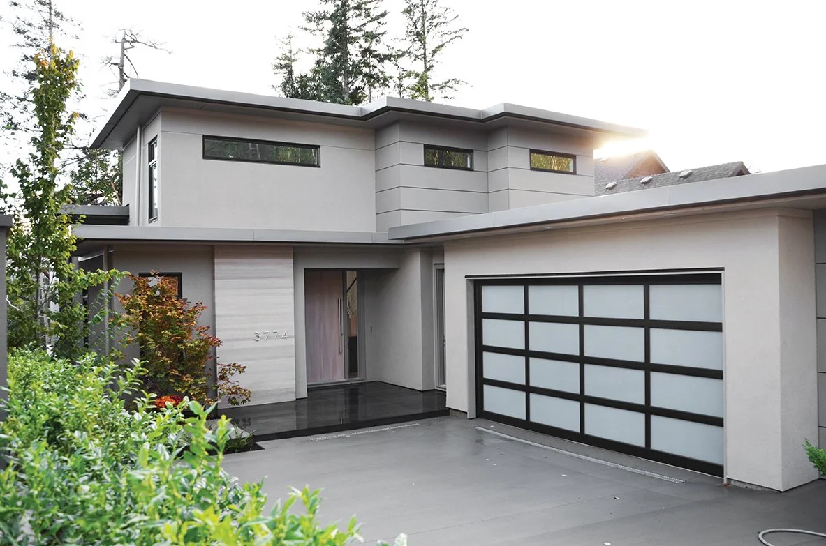 Modern two-story house with a sleek design, white exterior walls, large windows, and a black garage door, surrounded by greenery.