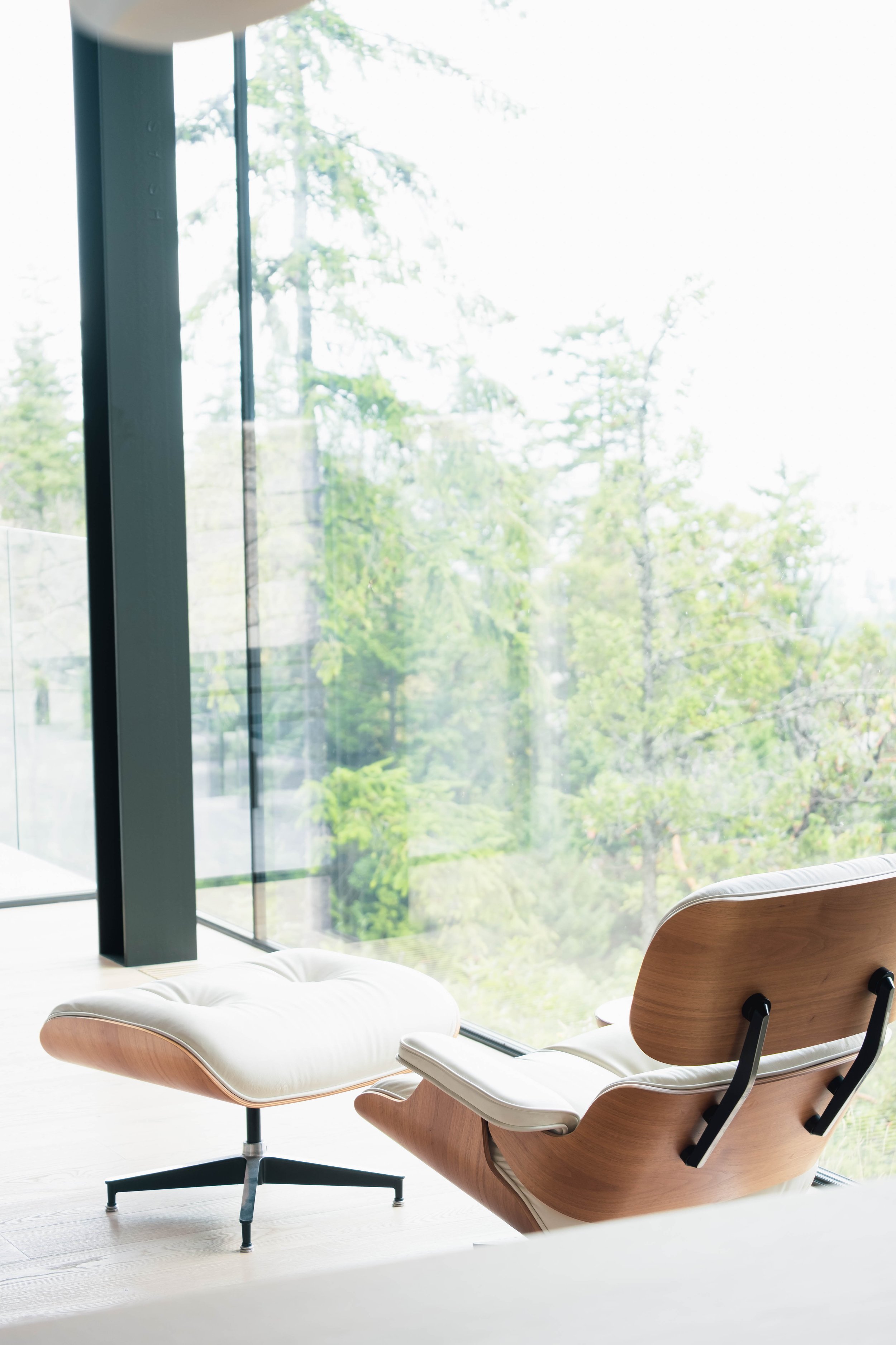 Modern wooden lounge chair with white cushion and matching ottoman in front of large window with a view of greenery outside.