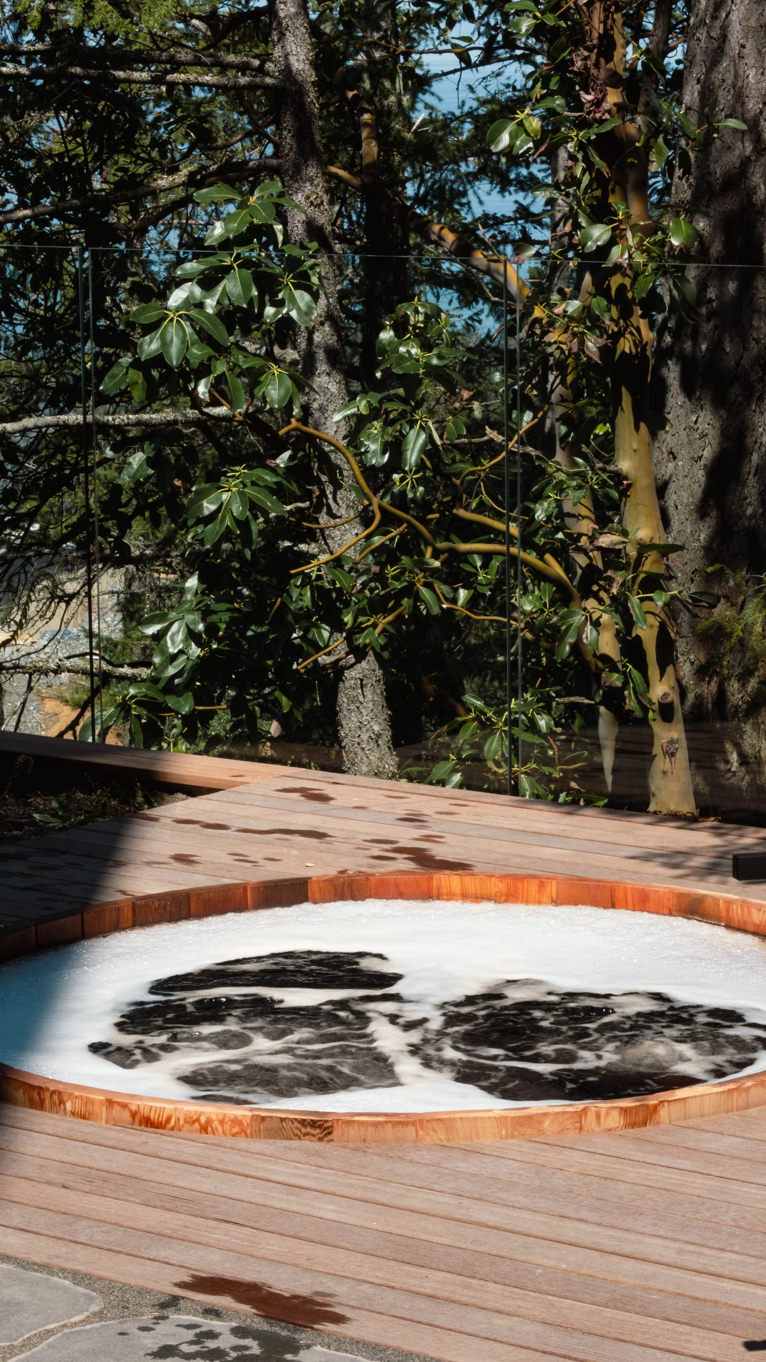 An outdoor hot tub with bubbling water on a wooden deck, surrounded by trees and a glass railing.