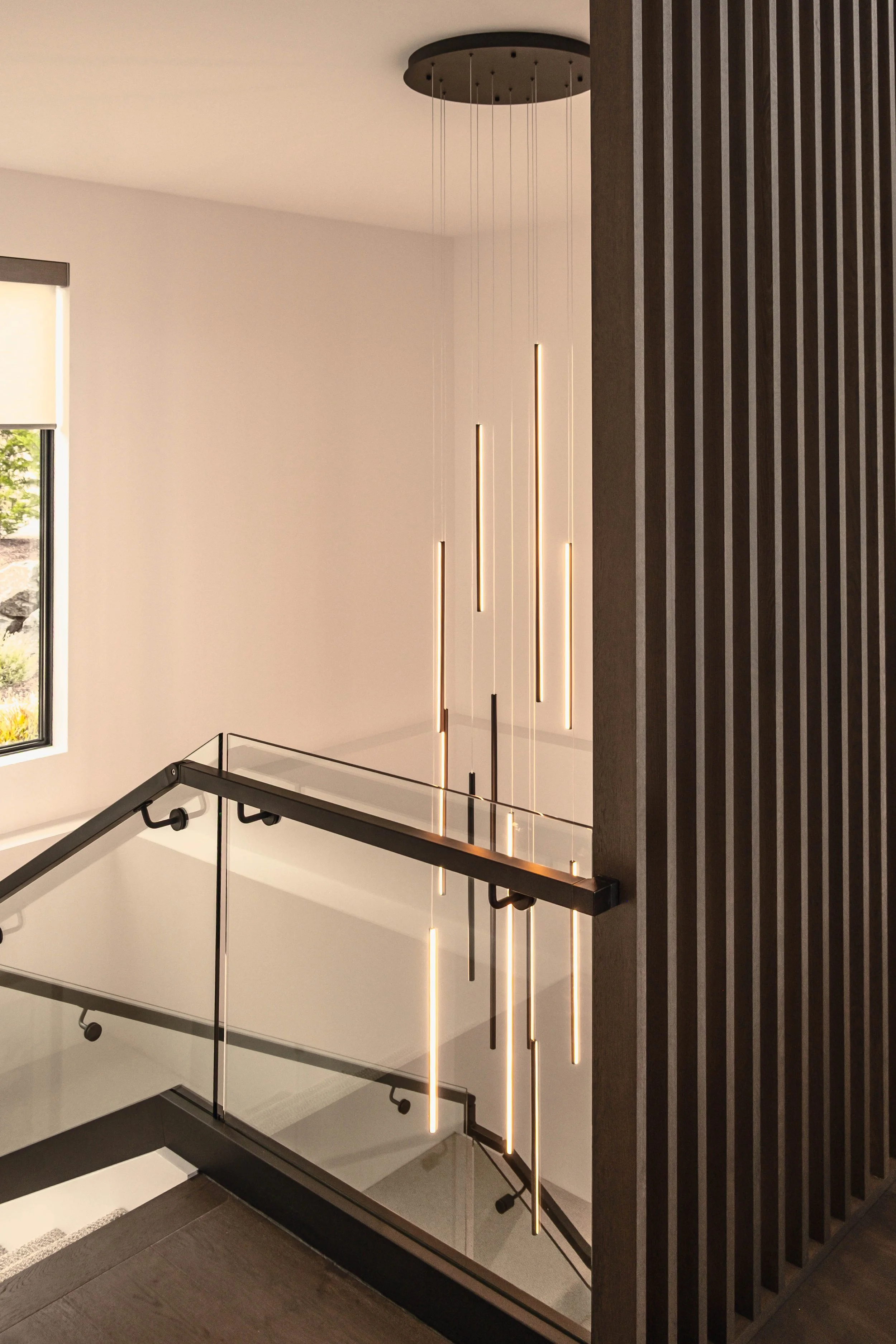 Modern indoor staircase with glass and black metal railing, next to a wall with vertical wooden slats, and unique hanging vertical LED light fixtures on the wall.