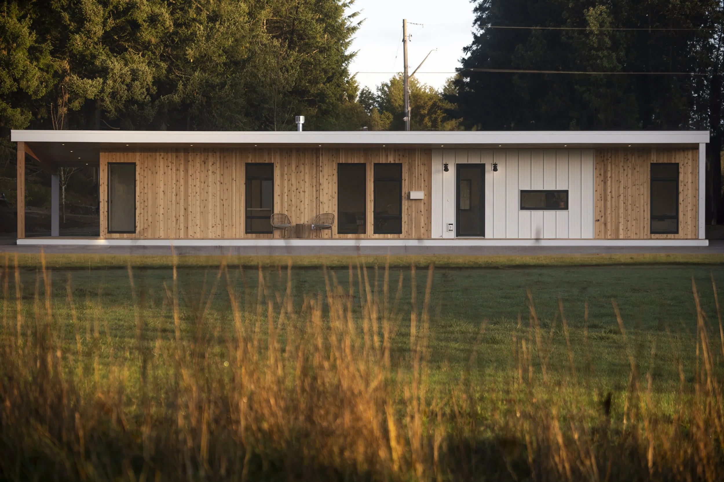 A modern, single-story house with wood and white siding exterior, large windows, and a covered porch with two chairs, set against a wooded backdrop.