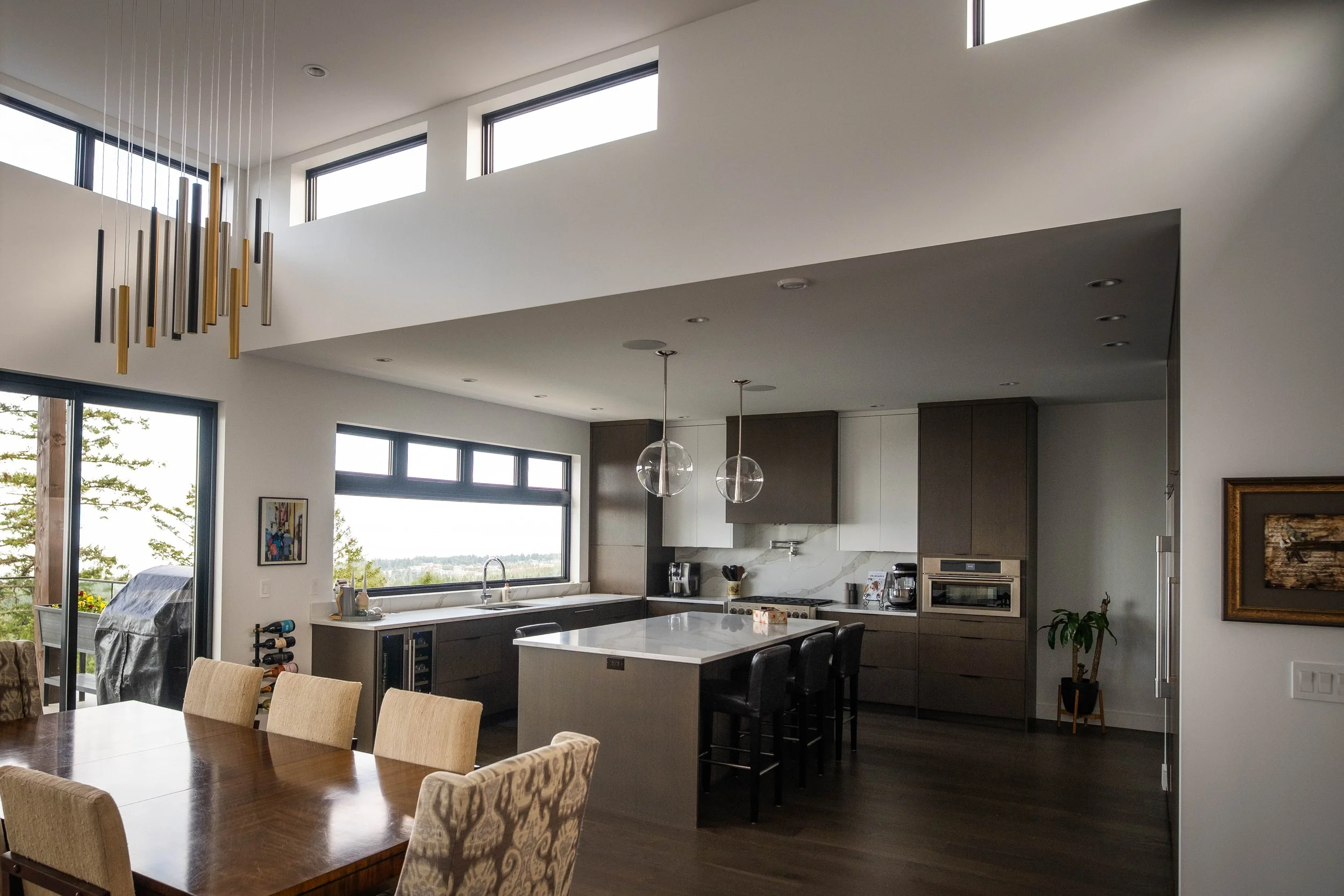 Modern open-concept kitchen with a white island, dark cabinets, large windows, and a dining area.