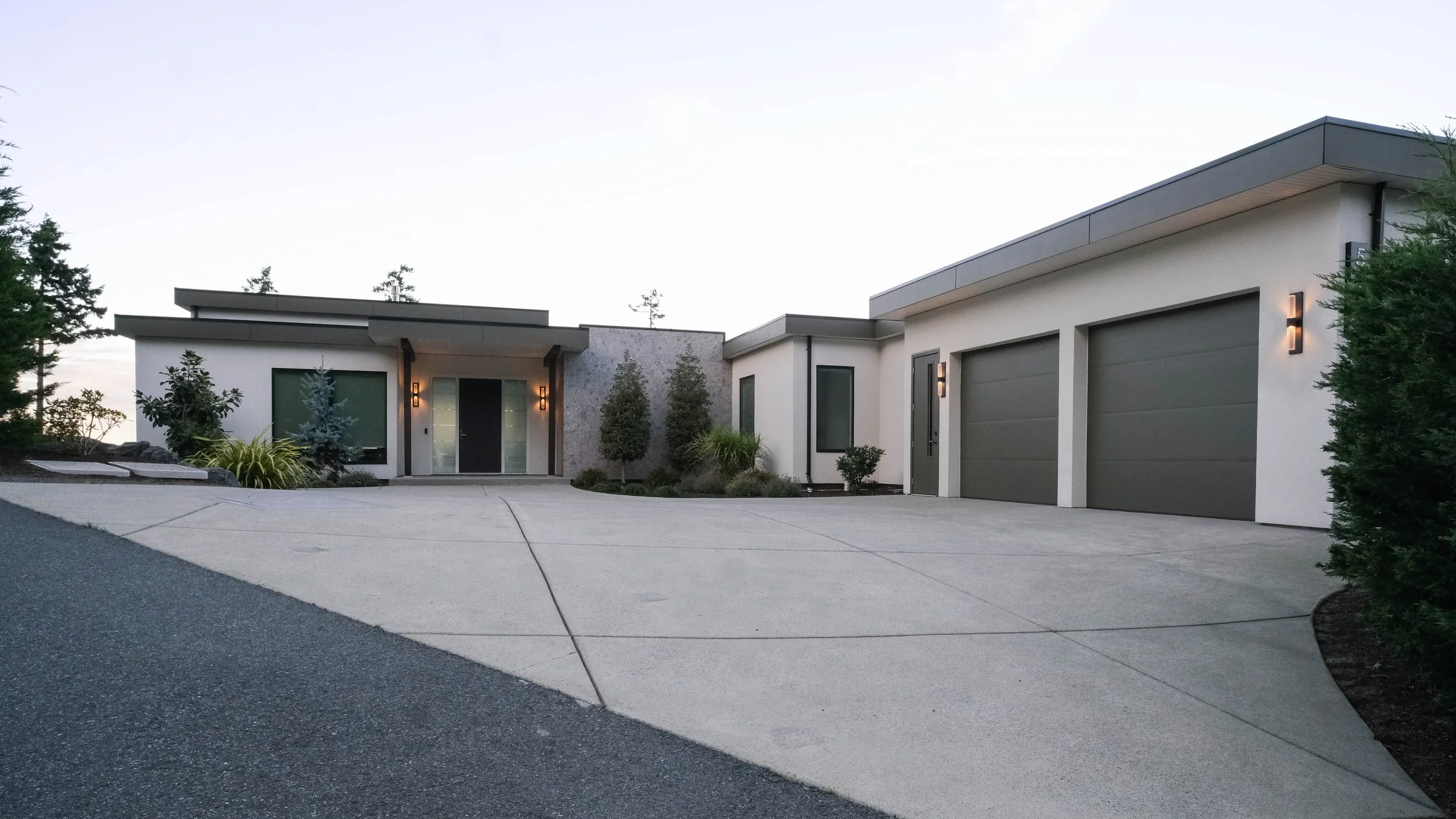 Modern house with a white exterior, flat roof, large windows, and an attached garage. The driveway is made of concrete, leading up to the garage and entrance, with landscaped plants and trees around the house.