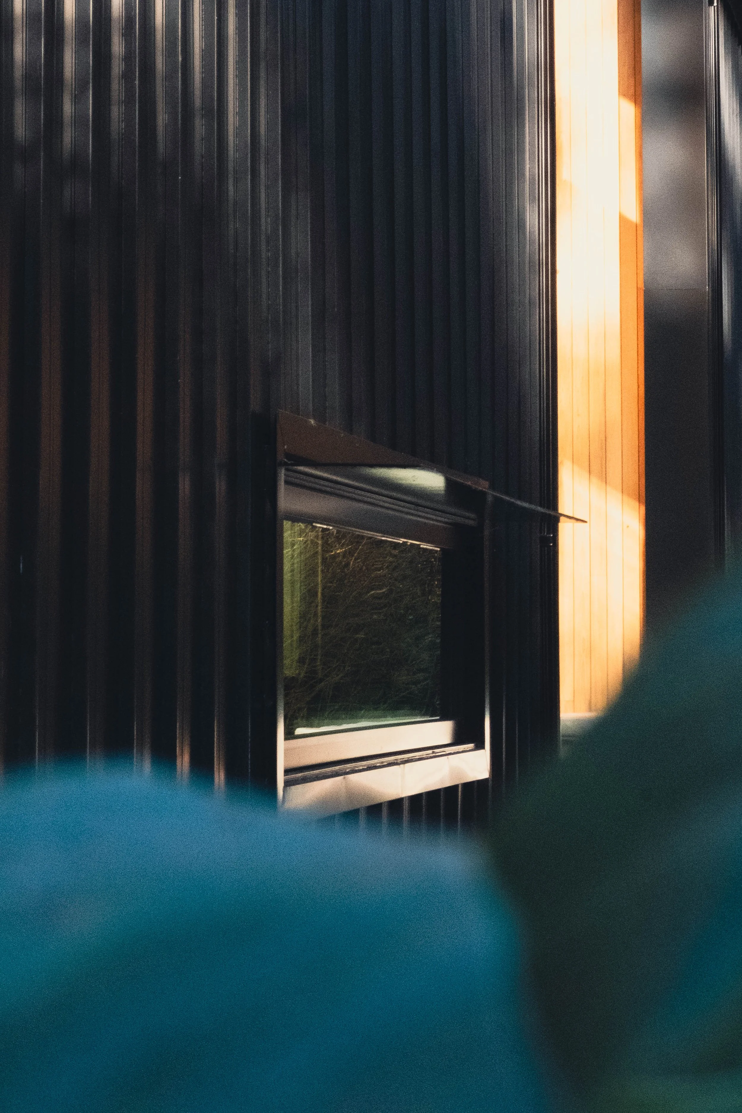 Close-up of a black, wooden exterior wall with a small, rectangular window and a sunlit wooden panel in the background.
