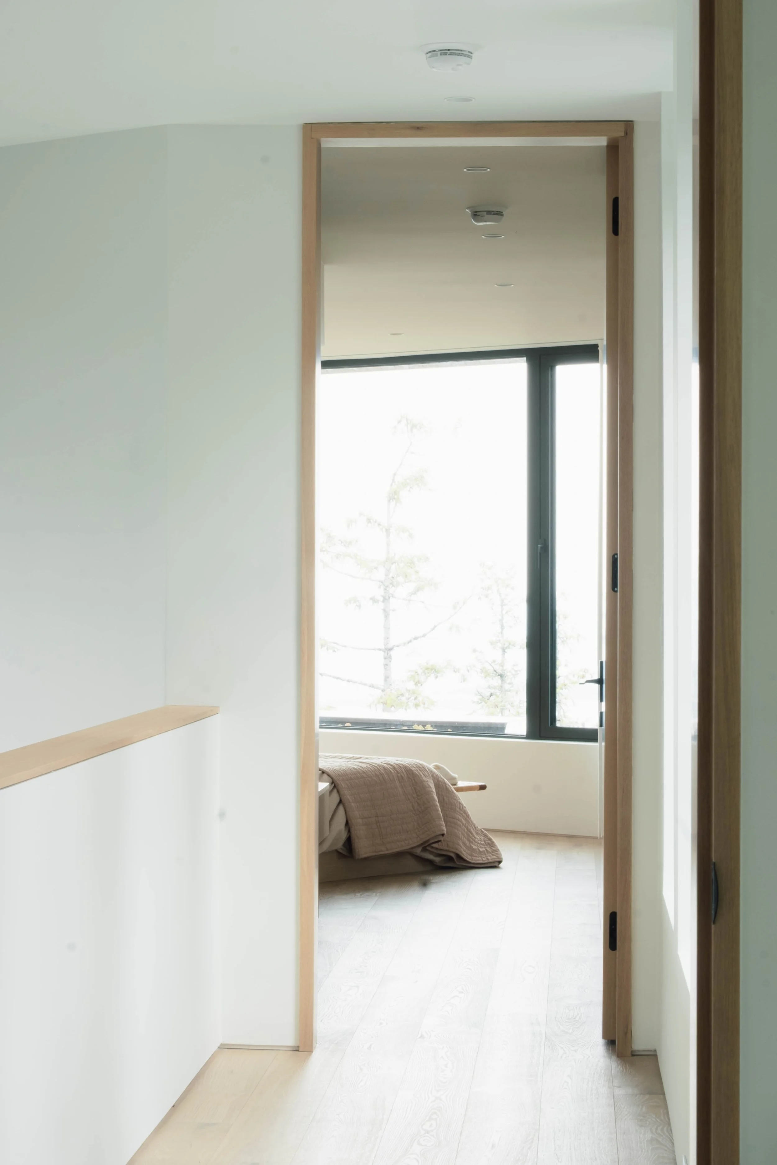 A minimalist bedroom with a large window showing a view of trees. The bed has neutral-colored bedding and is positioned near the window.