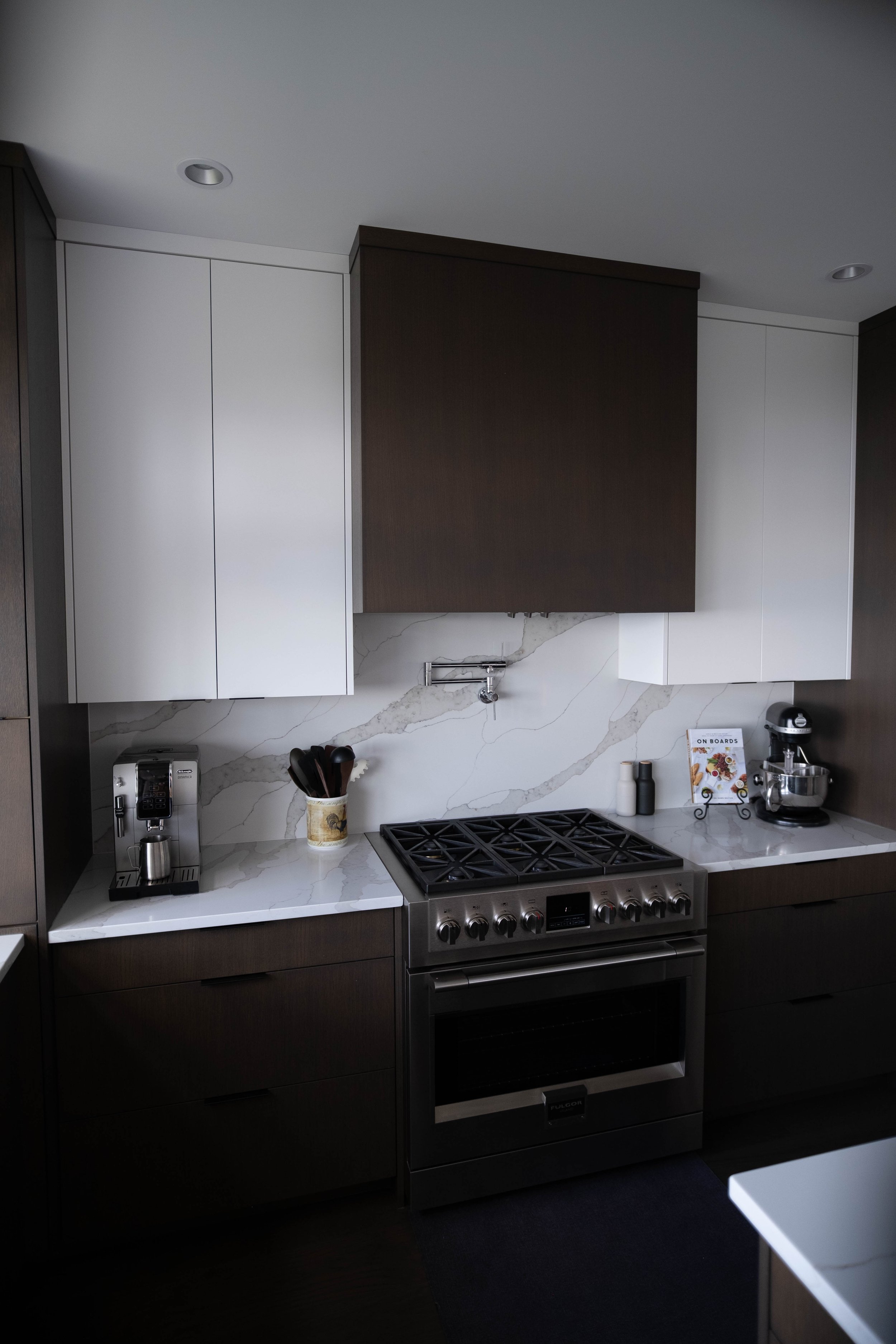 Modern kitchen with dark wood lower cabinets, white high cabinets, white marble backsplash, stainless steel oven, coffee maker, utensil holder, and stand mixer.