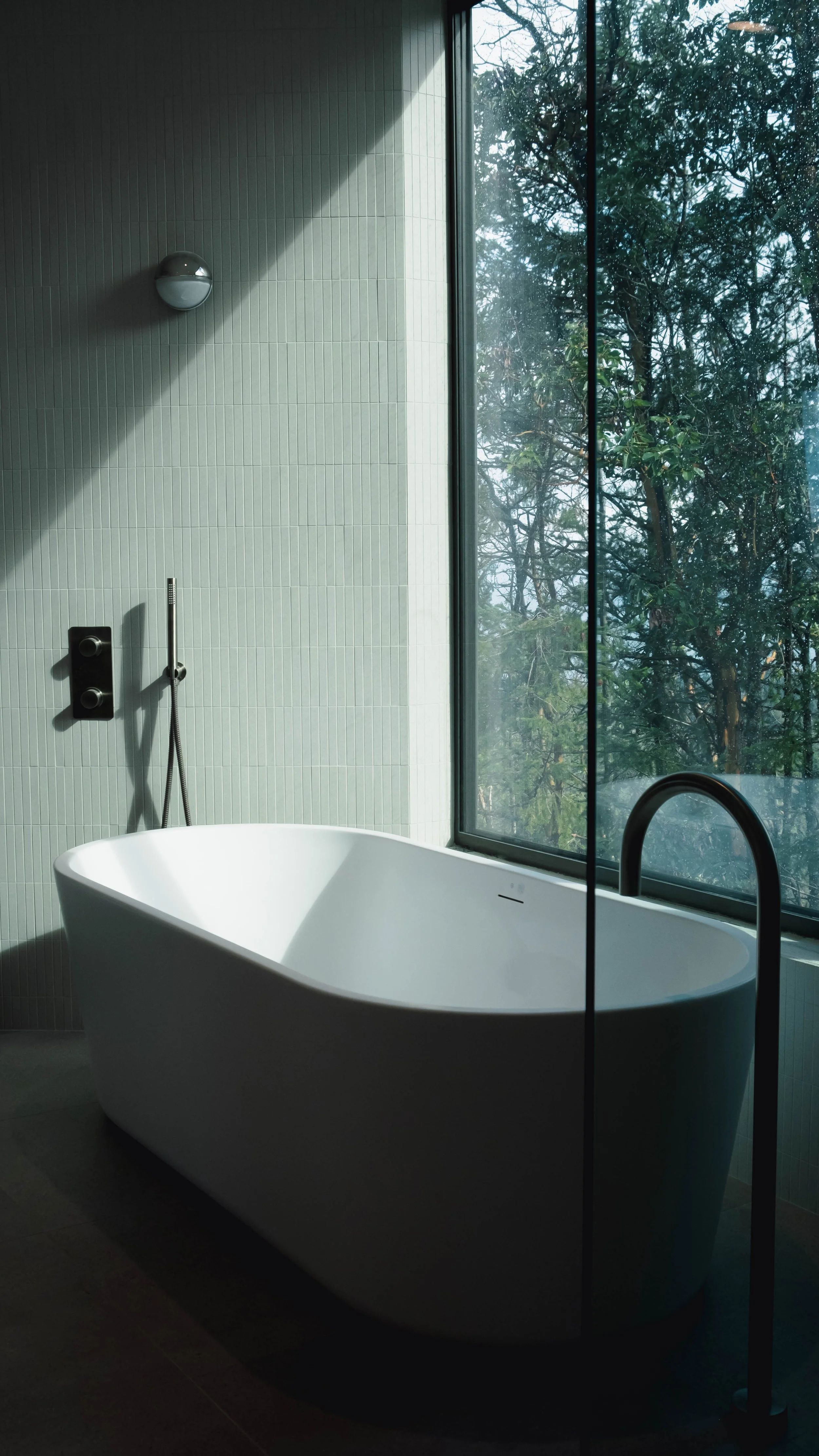 Modern bathroom with a white freestanding bathtub near a large window with trees outside, and a wall-mounted shower fixture.