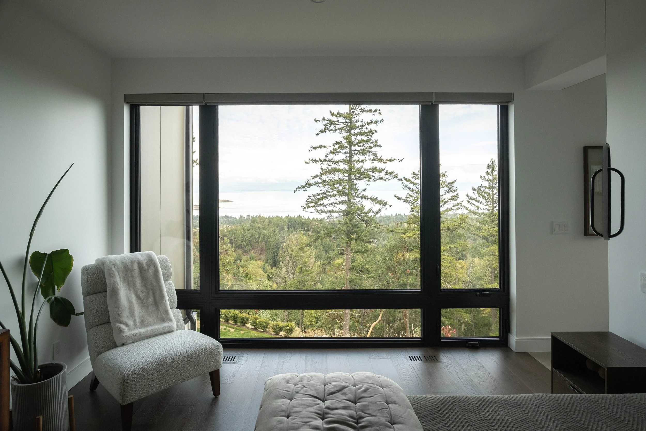 Living room with large window overlooking trees and nature, white armchair with throw blanket, potted plant, and minimal decor.