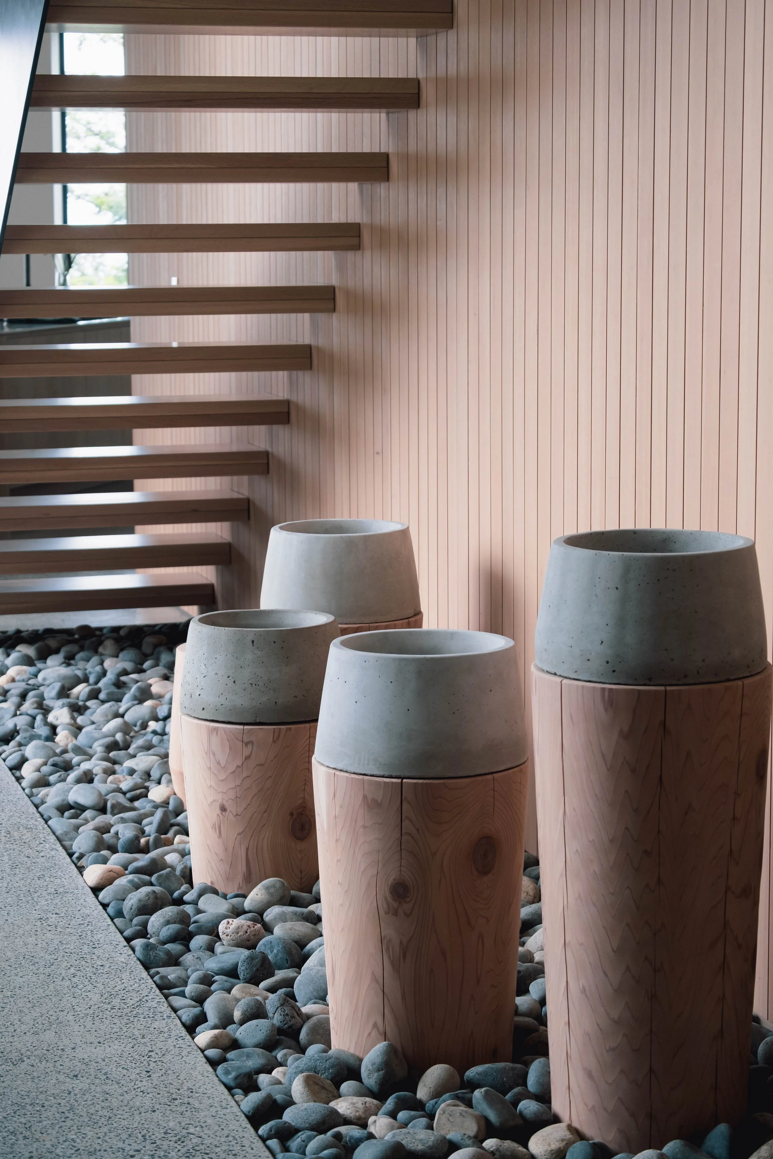 Four modern decorative stools with concrete tops and wooden bases are placed on a bed of small rocks, against a wooden wall and staircase in a contemporary interior.