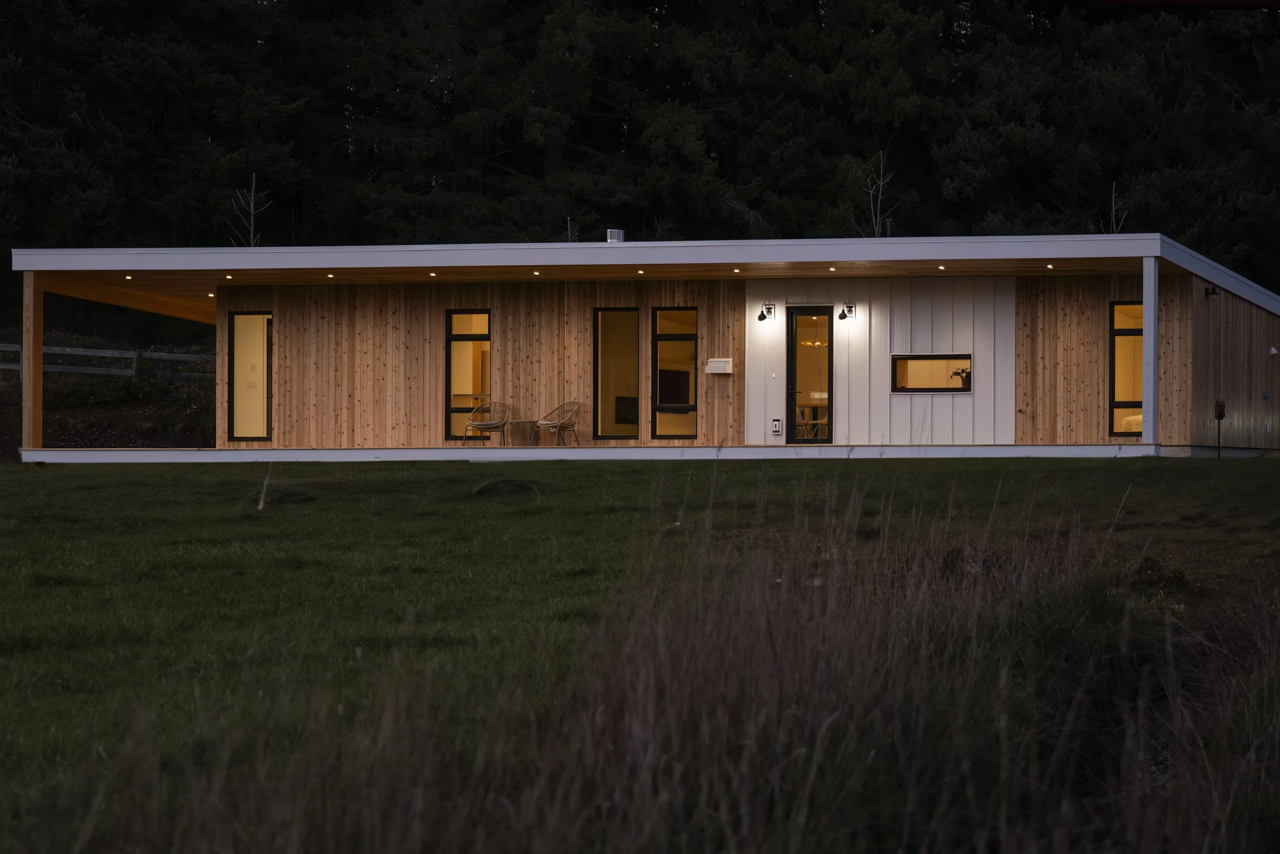Modern single-story house with wooden and white exterior walls, large windows, outdoor seating, lit interior, set in a grassy area at dusk.