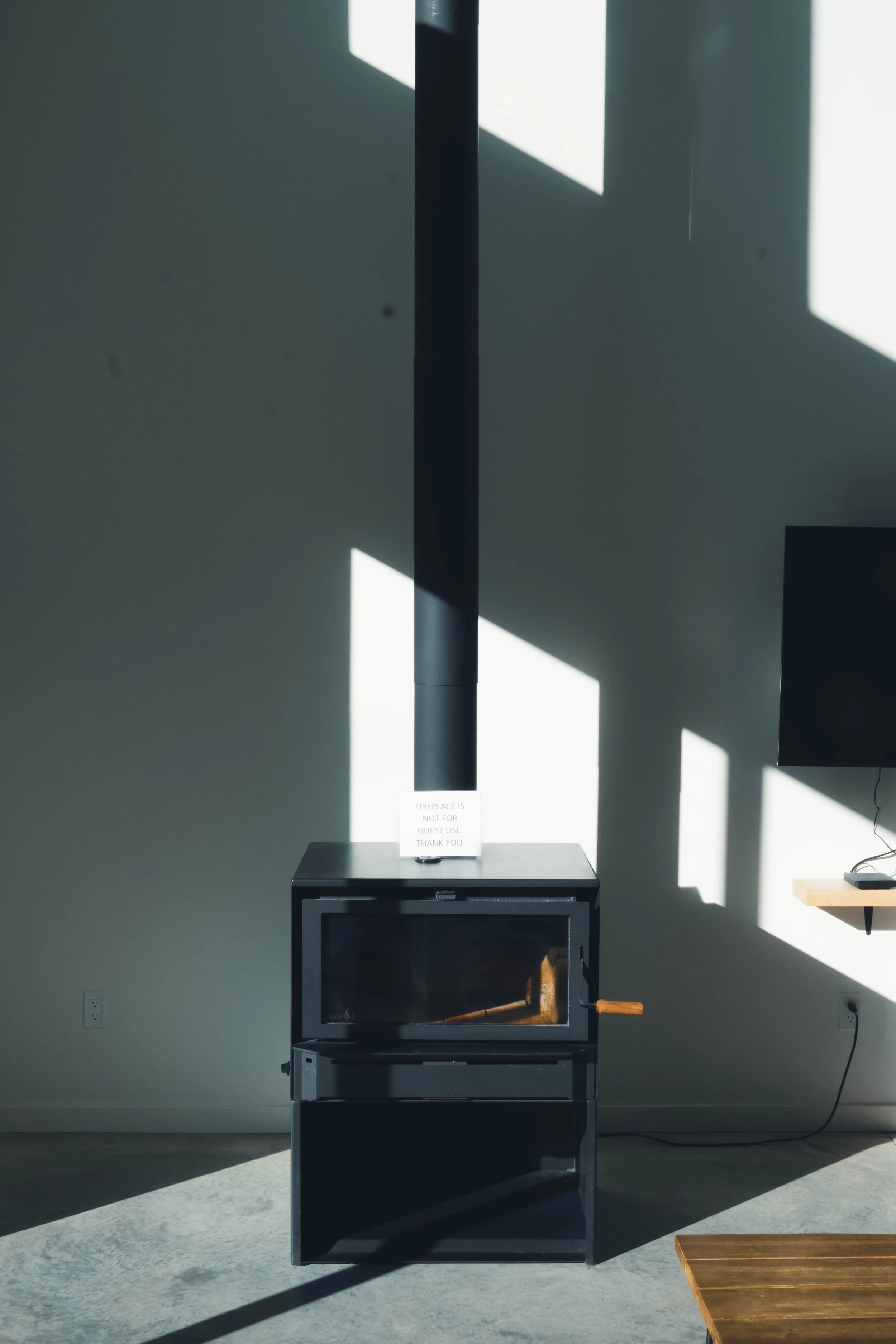A black wood stove with a metal chimney pipe against a wall, with sunlight casting geometric shadows on the wall and floor.