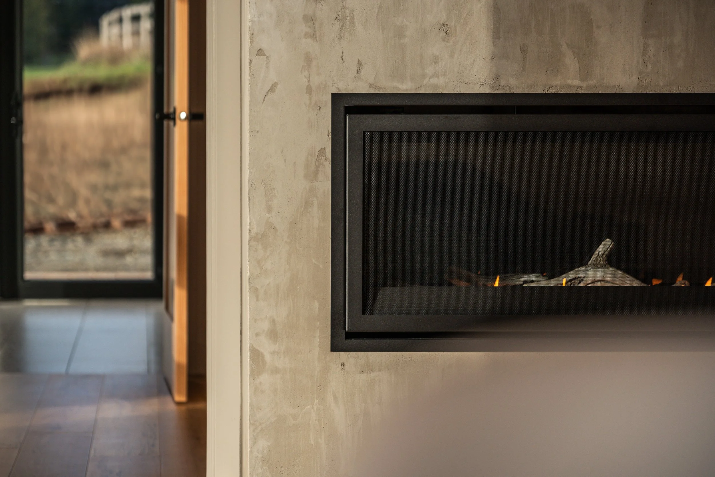 Close-up of a modern fireplace with visible logs, set within a concrete wall, with an open door or window showing an outdoor landscape in the background.
