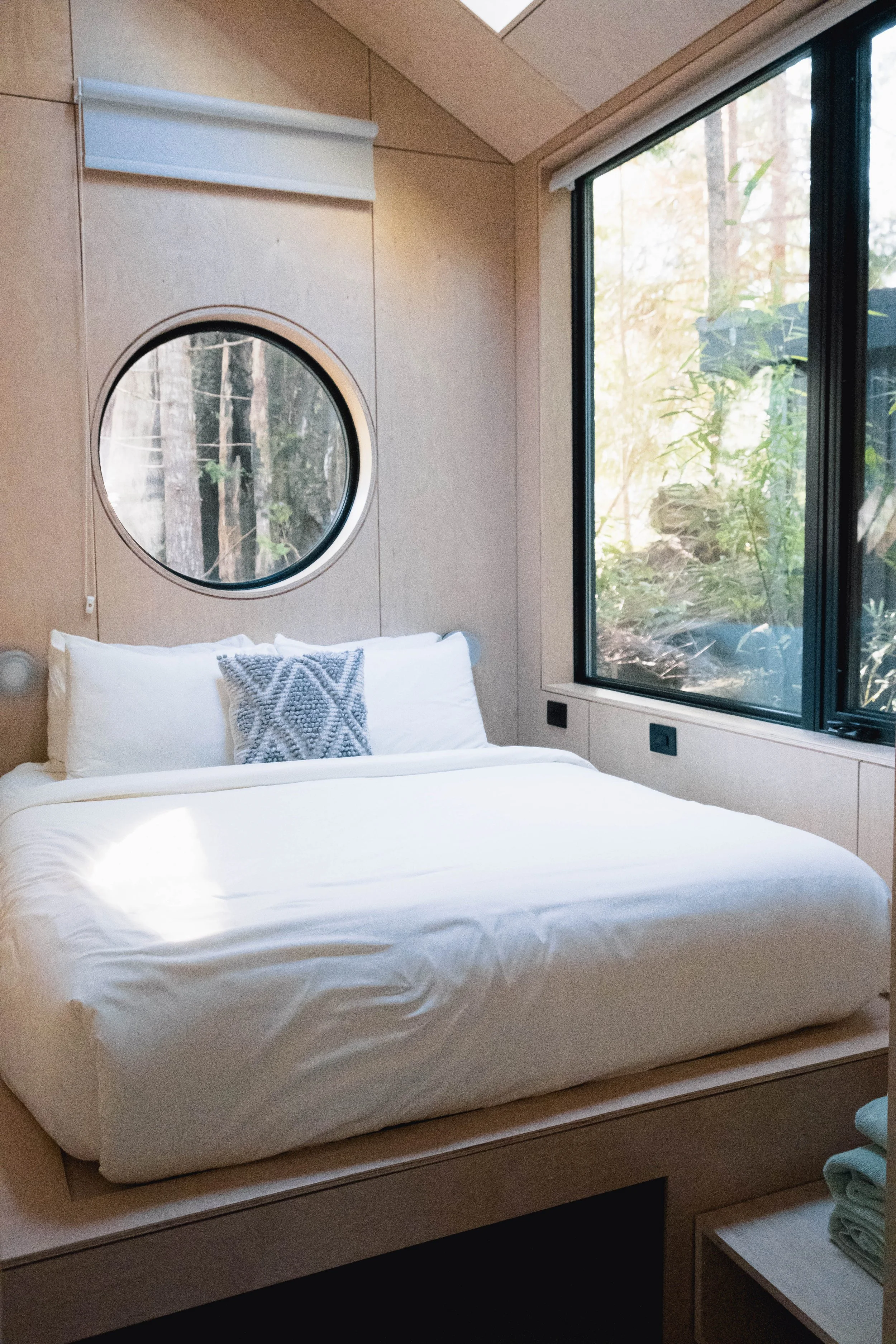 A cozy bedroom features a neatly made bed with white bedding and a decorative pillow. There is a large window on the right side showing a forest view, and a round mirror on the wall above the bed.