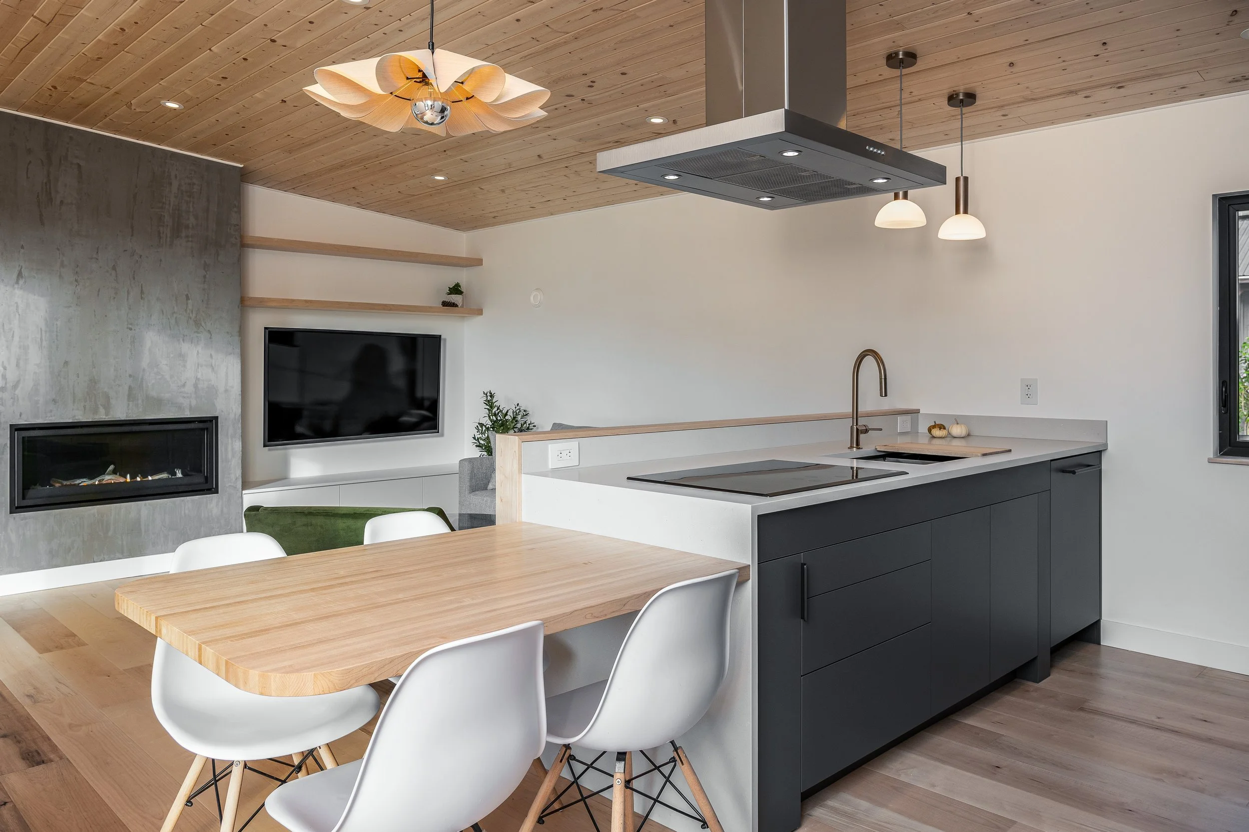 Open-concept kitchen with black lower cabinets, white countertop, modern faucet, and stovetop, adjacent to a dining area with a light wood table and white chairs. A living area in the background features a gray wall, mounted flat-screen TV, and a sle