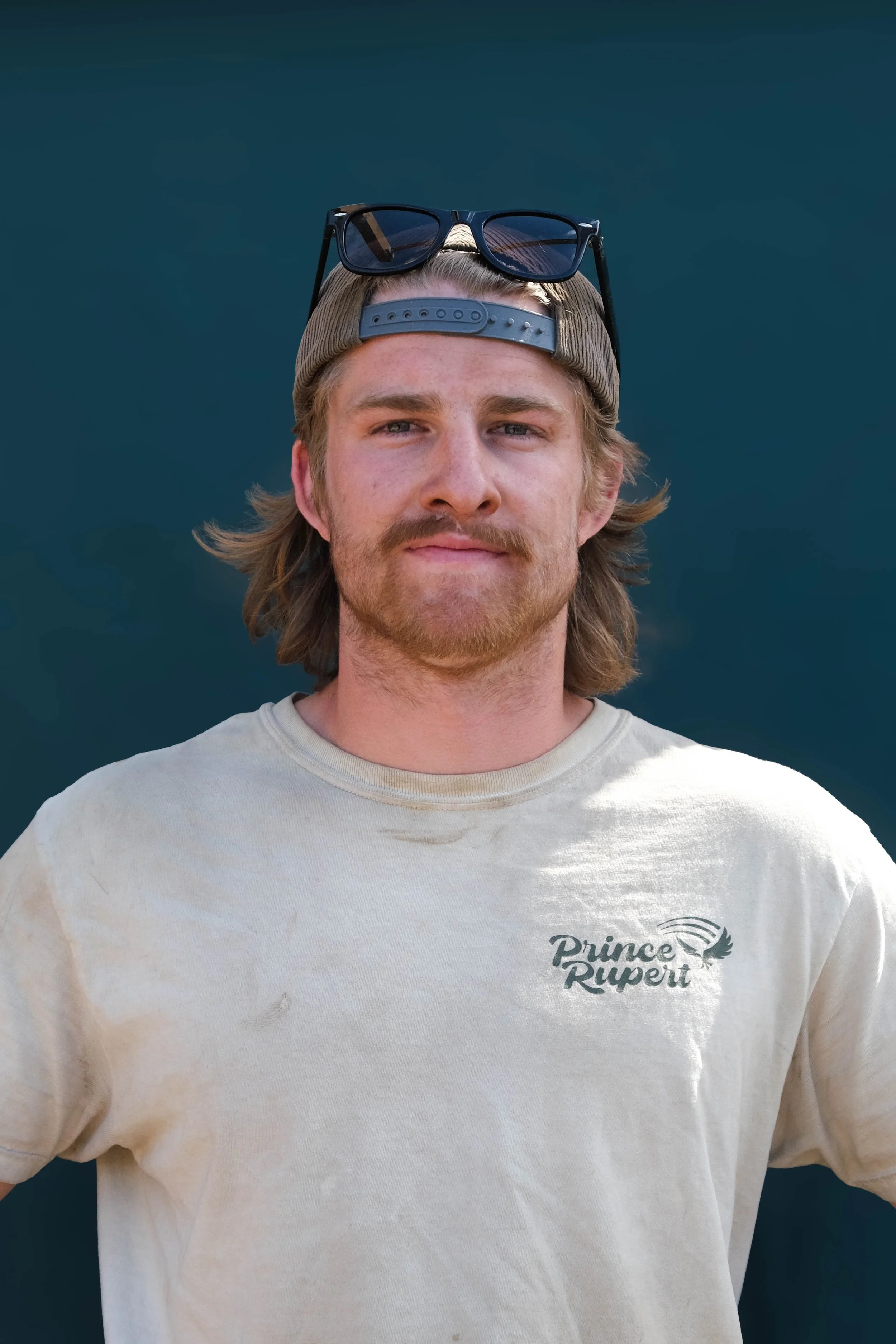 A young man with long hair, a beard, and sunglasses on his cap, wearing a beige Prince Rupert T-shirt, stands against a dark background.
