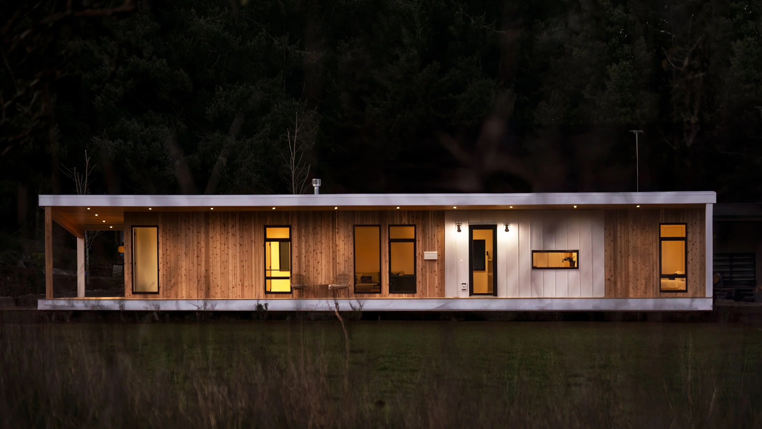 Modern, single-story house with wooden and white exterior panels, large windows, and outdoor seating, illuminated at dusk and reflecting on a pond in front.