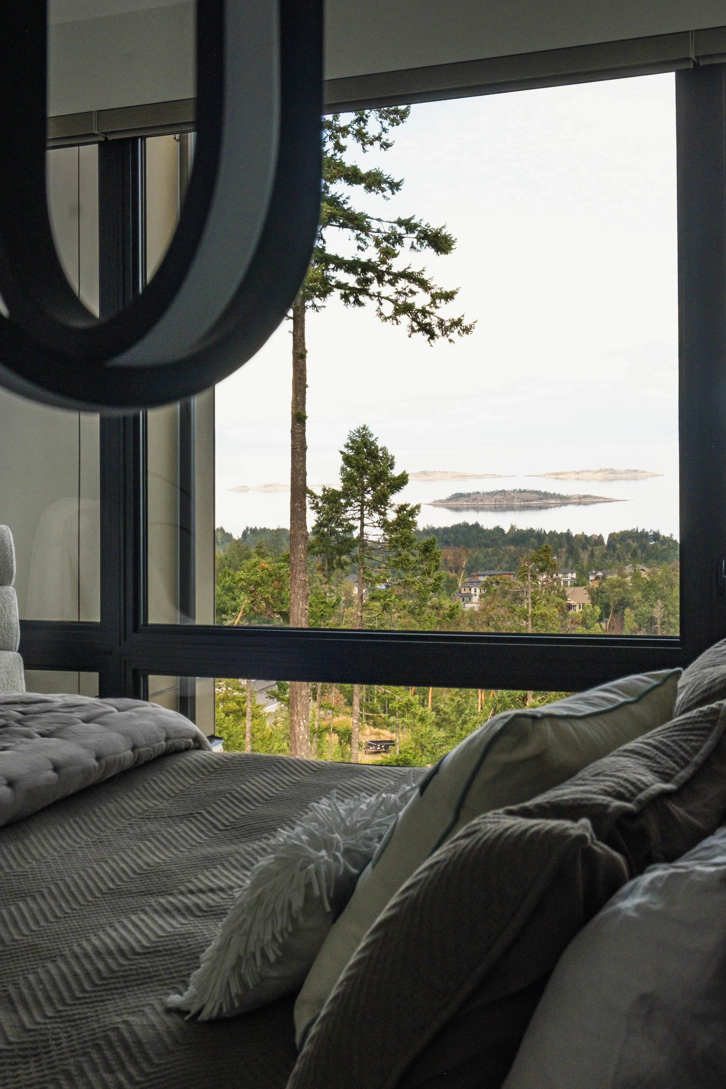 A bedroom with a large window showing a view of trees, hills, and water in the distance.