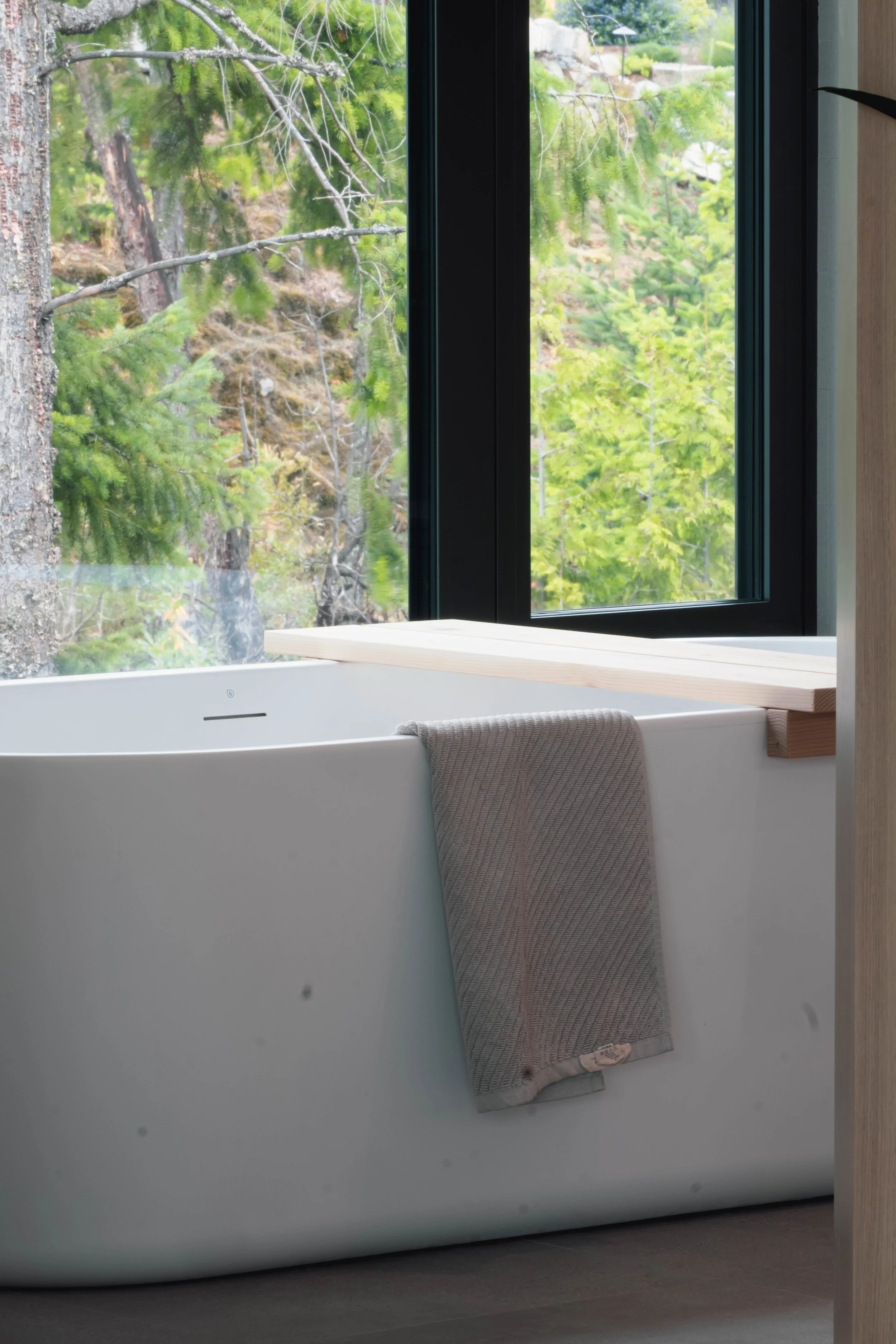 A modern bathtub near a large window with a view of green trees and foliage outside. A towel hangs over the edge of the tub.