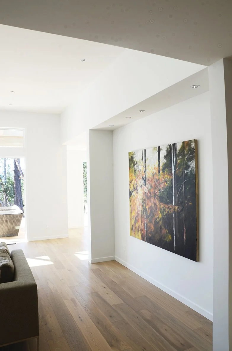 A modern living room with white walls, wooden flooring, and a large landscape painting of trees with autumn foliage on the wall. Sunlight streams in through a sliding glass door, revealing an outdoor space with trees.